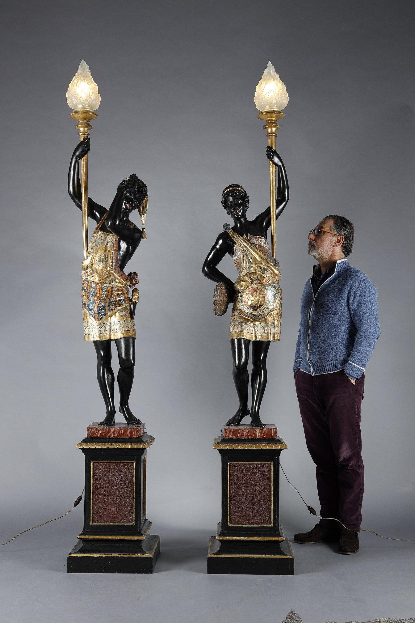 Couple of Nubian torchbearers in carved wood with black lacquered bodies and richly decorated with polychrome and gilded garments, standing on a square wooden base painted in imitation of porphyry marble. Electrically mounted with a polished glass