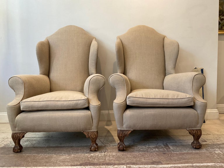 A large scale pair of upholstered English wing chairs dating from the 1920s.
These impressive chairs are upholstered in neutral slightly coarse linen with feather cushions and oak carved ball and claw feet.

A great functional and comfortable