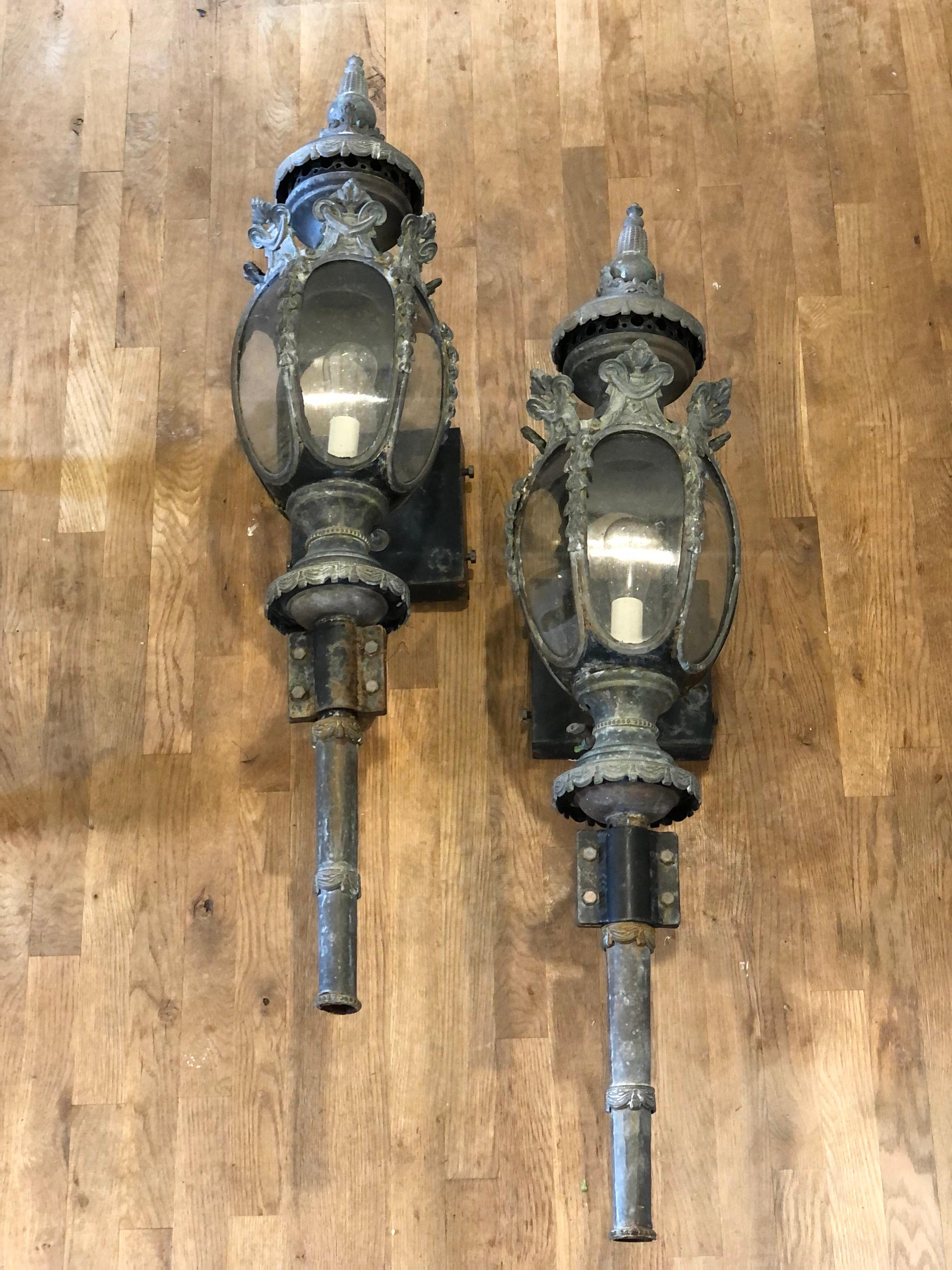 Pair of large lanterns with a mix of bronze, brass and iron. 
Tapered lanterns surmounted by a flame echoing their past as gas lanterns. Have been wired for electricity but could be converted back to gas. 
Curved glass. Bronze and brass has