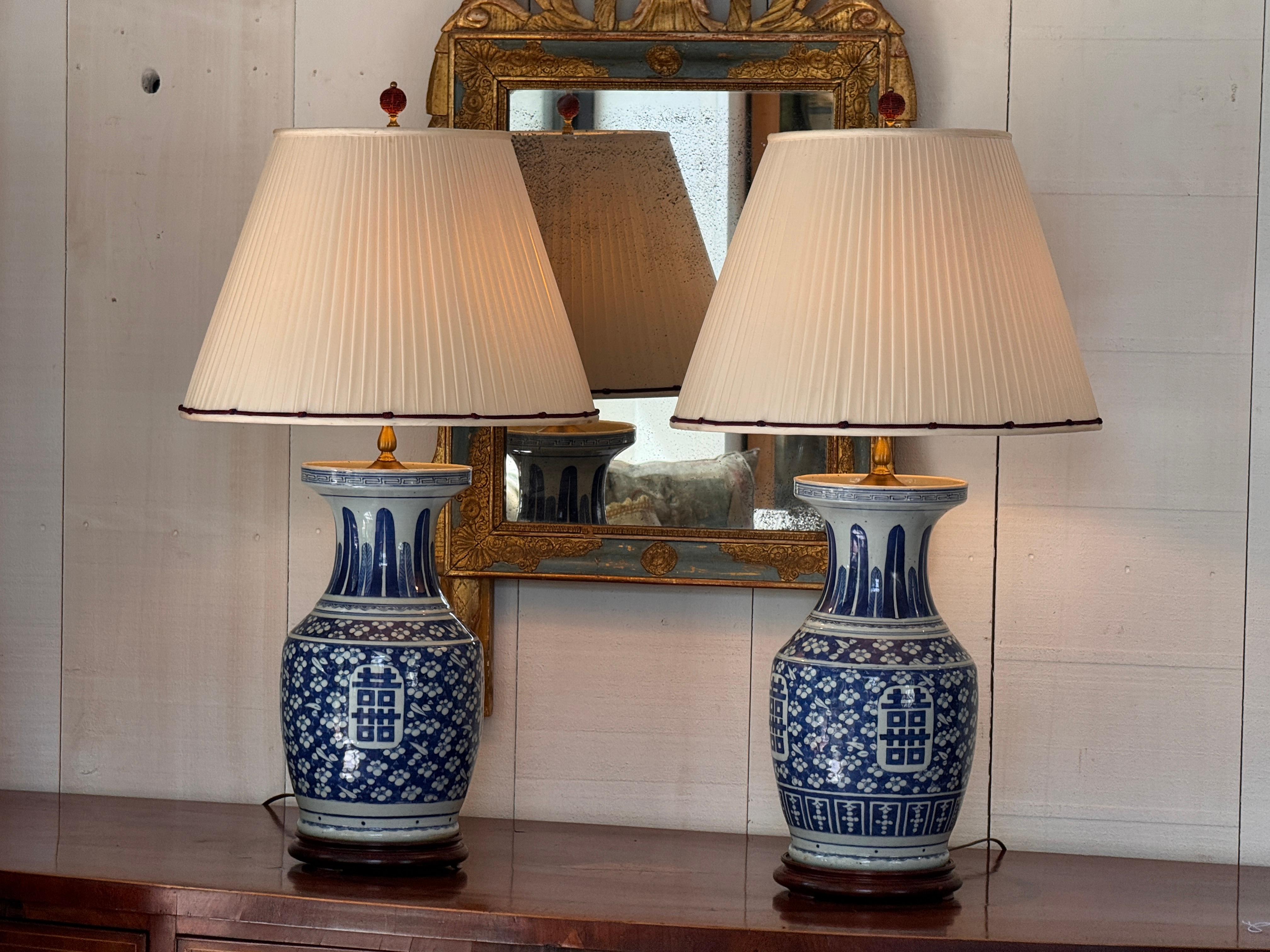 Coppia di grandi vasi in porcellana bianca e blu montati come lampade, decorati con spruzzi floreali e riserve di personaggi di buon auspicio. I corpi si assottigliano fino a raggiungere i colli dipinti con pannelli verticali di foglie stilizzate,