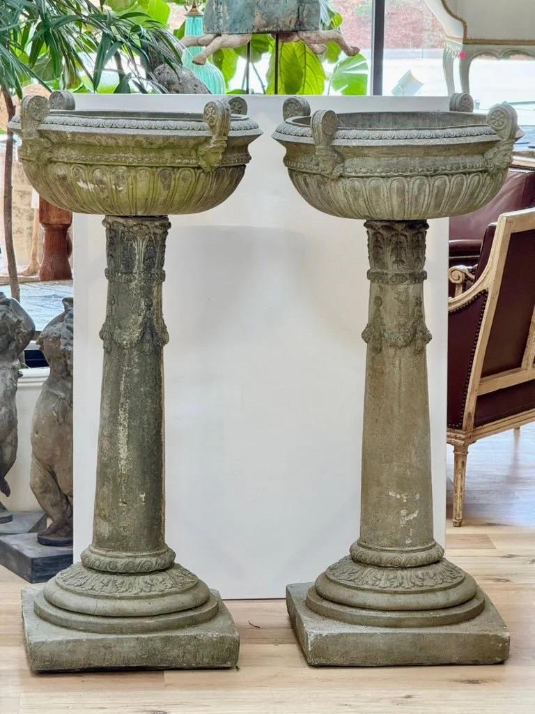 Pair of Large Cast Stone Urns having egg-and-dart rim with four female masks and lobed bowl, on a round pedestal base with acanthus leaf, laurel garland, and guilloche designs in relief. 49 x 24 x 24 in