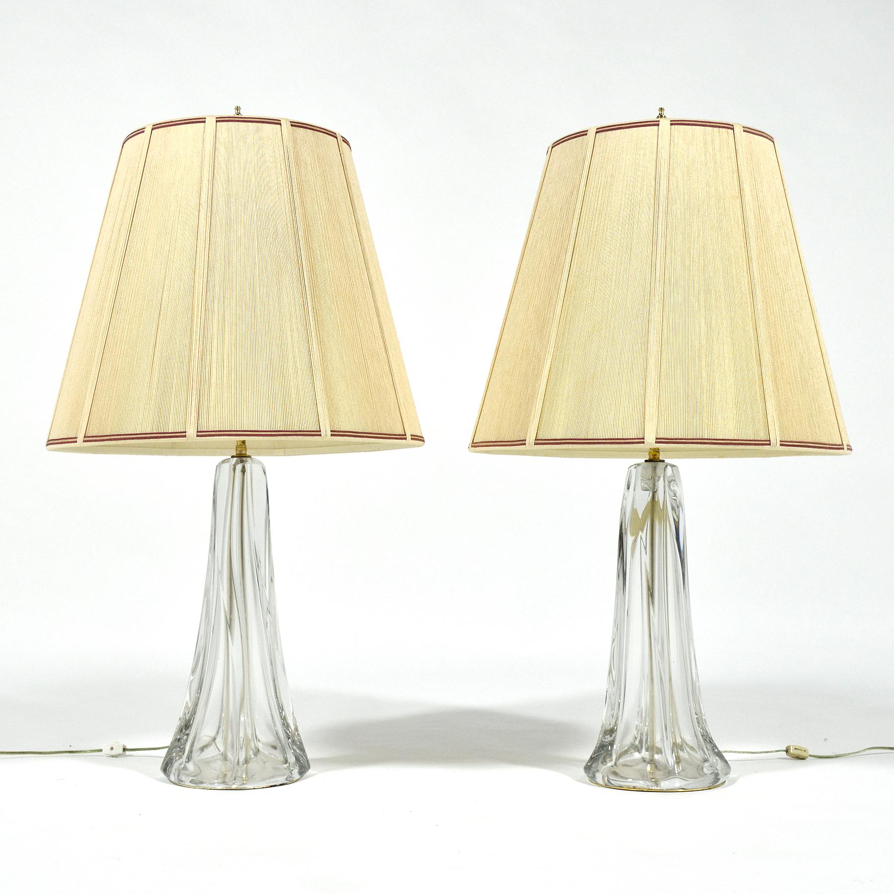 This pair of large crystal table lamps are unmarked, but are very similar to works by Val St Lambert. The substantial clear bases have a swirled form and support their original handmade string shades.