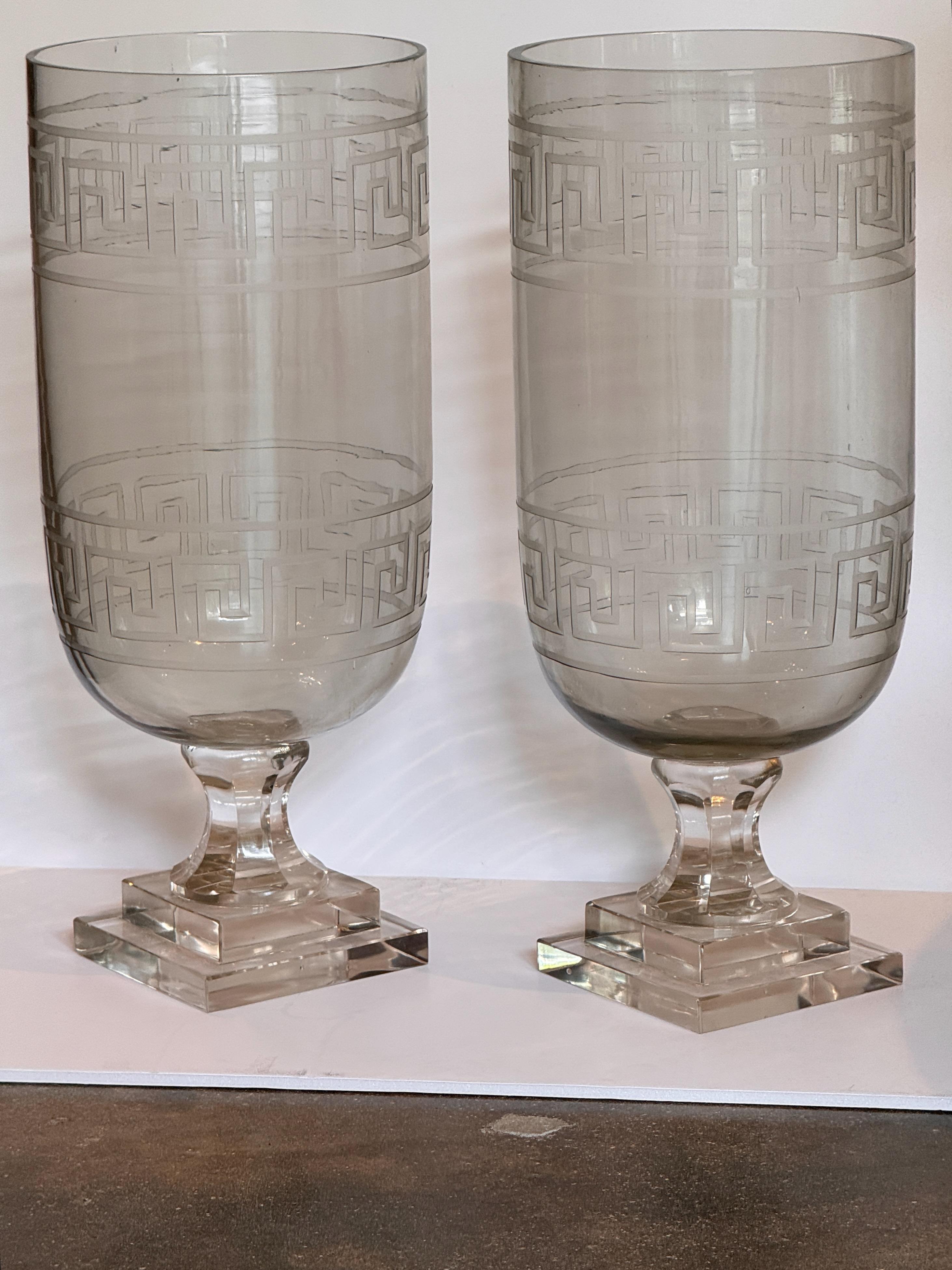 Tall clear glass vases etched with a bold Greek key band, each raised on a tiered square base. Crisp geometric lines contrast with the rounded bowl form, creating a clean neoclassical statement.