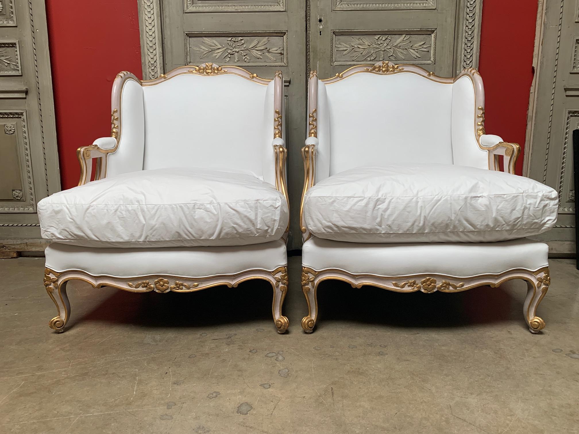 A pair of carved wood French Louis XV style armchairs, bergeres with a painted pink and gilded finish.
These large chairs are hand carved with wings and raised floral carving. They are upholstered in muslin and have a large down and feather cushion.