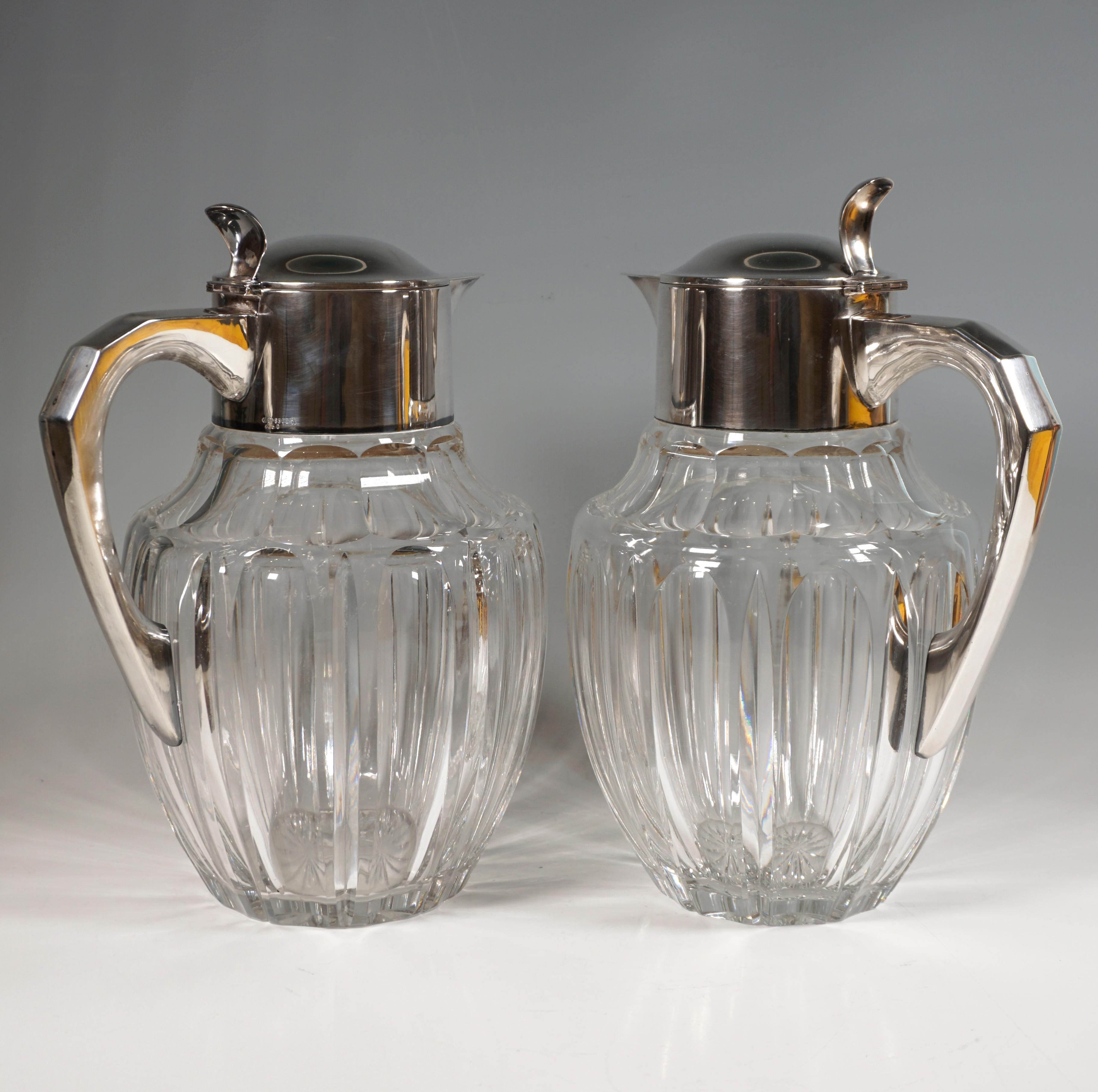 A pair of voluminous, bulbous, cut clear glass carafes on a round base, plain, smooth silver mountings with hinged lids with thumb rests and J-shaped handles with beveled accents on the outside.

Dimensions:
height: 31.0 cm / 12.20 in
width: