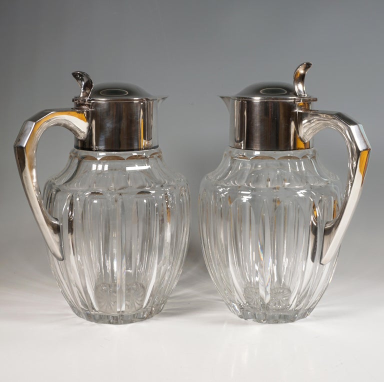 A pair of voluminous, bulbous, cut clear glass carafes on a round base, plain, smooth silver mountings with hinged lids with thumb rests and J-shaped handles with beveled accents on the outside.

Dimensions:
height: 31.0 cm / 12.20 in
width: