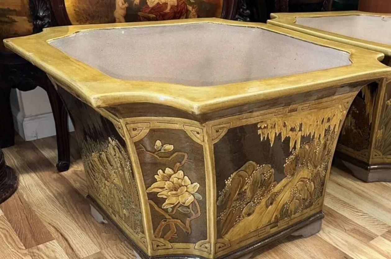 Una coppia di grandi fioriere in ceramica smaltata con un  Scena asiatica.  Le fioriere in terracotta vintage sono di colore ocra e marrone e presentano angoli dentellati e lati affusolati.  Le fioriere smaltate presentano fiori e una scena di