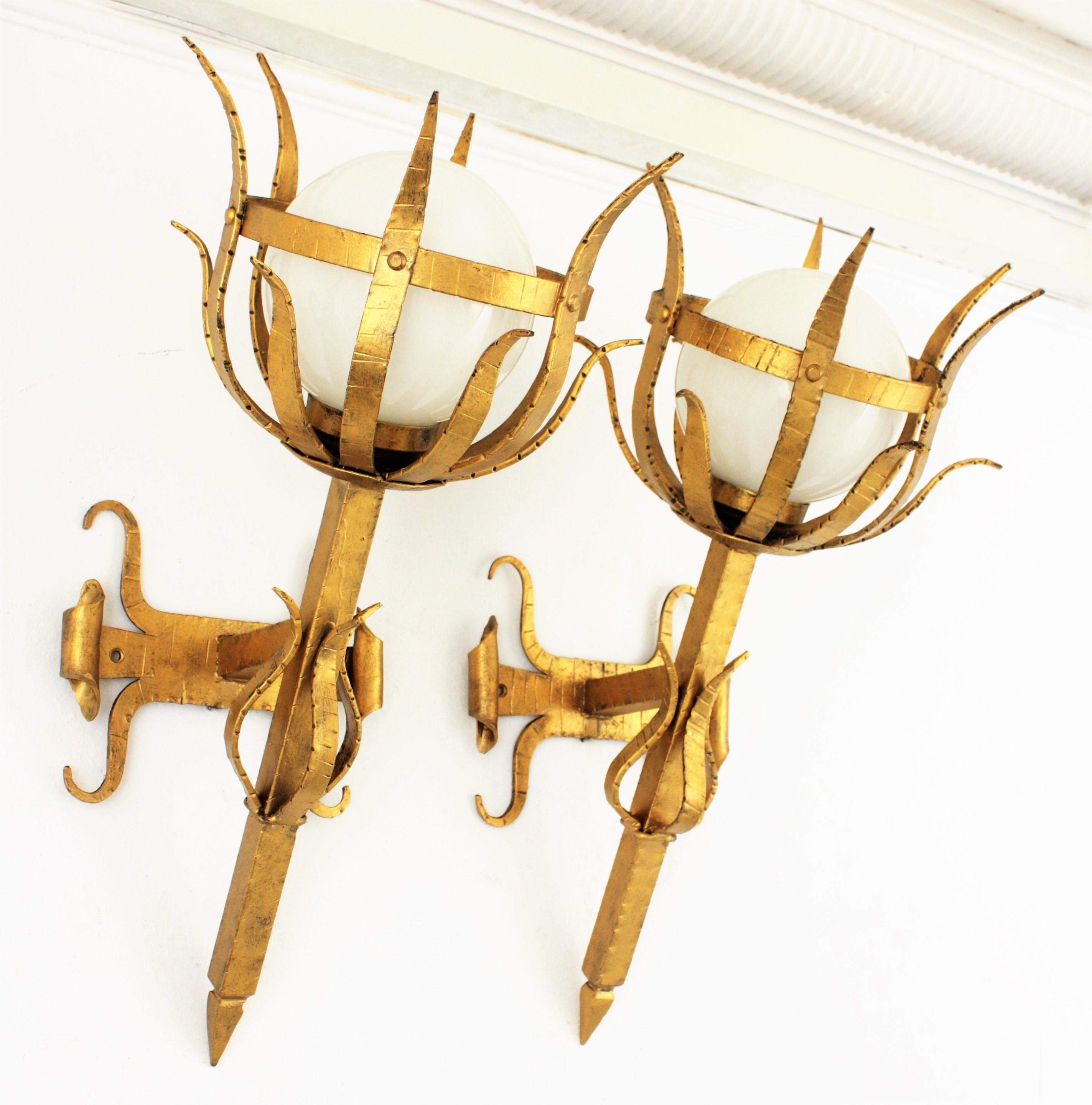 A huge hand-hammered iron Gothic style torch wall sconces with opaline glass globe shades and gilt patina finish,
Spain, 1950s.
This beautiful pair of wall lights can be placed indoor or outdoor (wired for both uses).