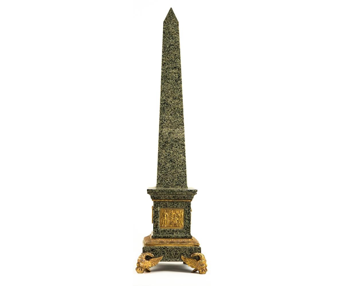 Offered is this fabulous pair of green, grey, black speckled granite obelisks with the obelisk form rising from a rectangular base decorated on one side with gold gilt bronze plaque featuring soldiers and a seated authority figure. The remains three