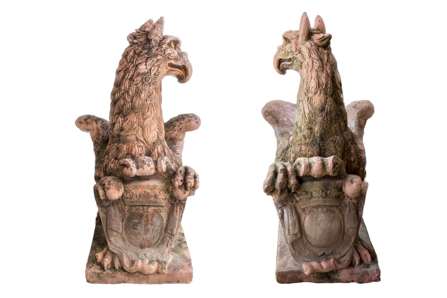 Pair of large griffons in reconstituted stone in terracotta manner. They have lion paws and tail, eagle wings and head with horse ears. They are in squat position on they back legs, folded wings along their back, turned head on side et front legs