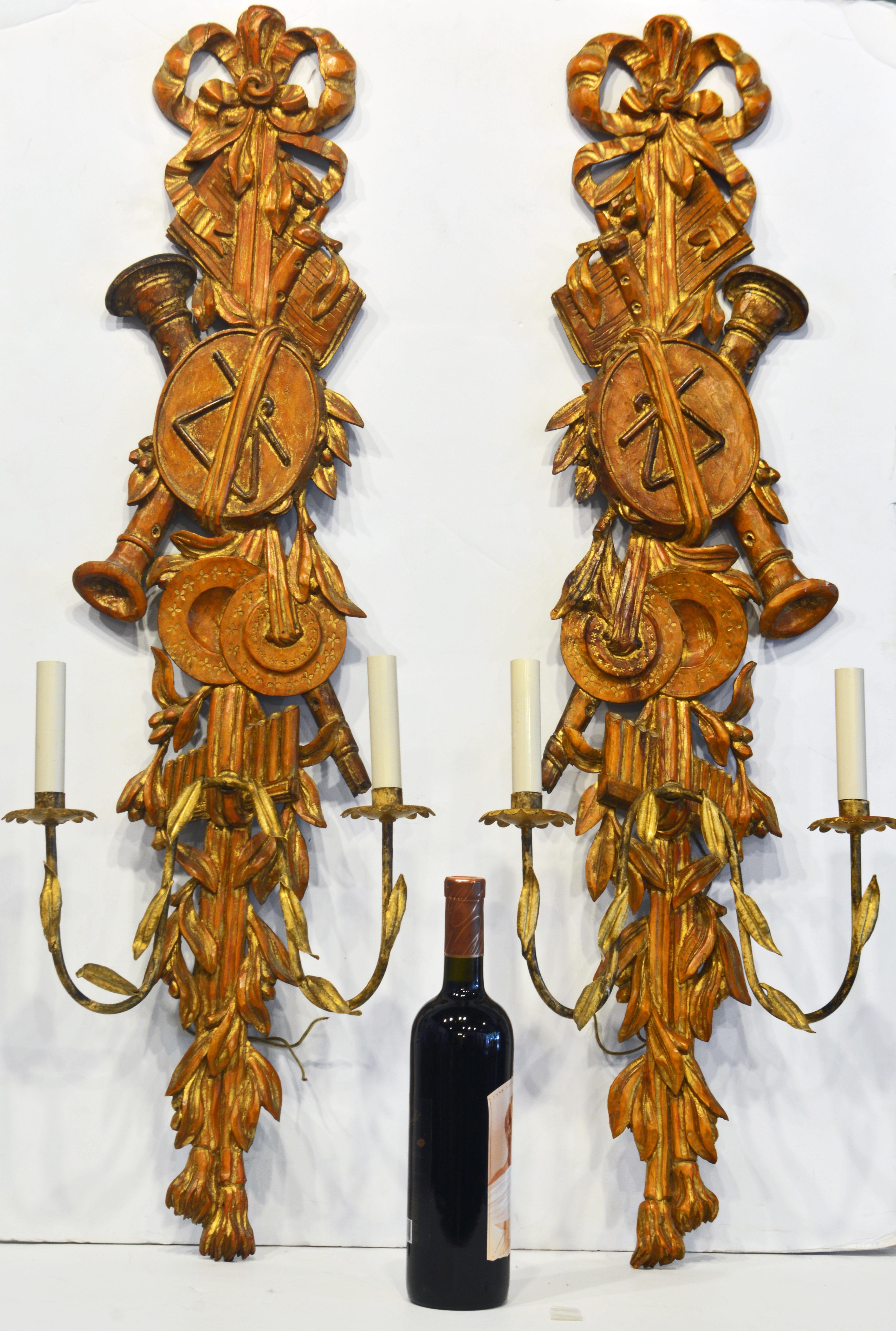 Almost 40 inches tall these Italian carved gilt and wood wall sconces are impressive. The carvings reflect musical instruments, leaf work, ribbons and berries. The two curved gilt metal light light arms are decorated with gilt tole leaves. The style