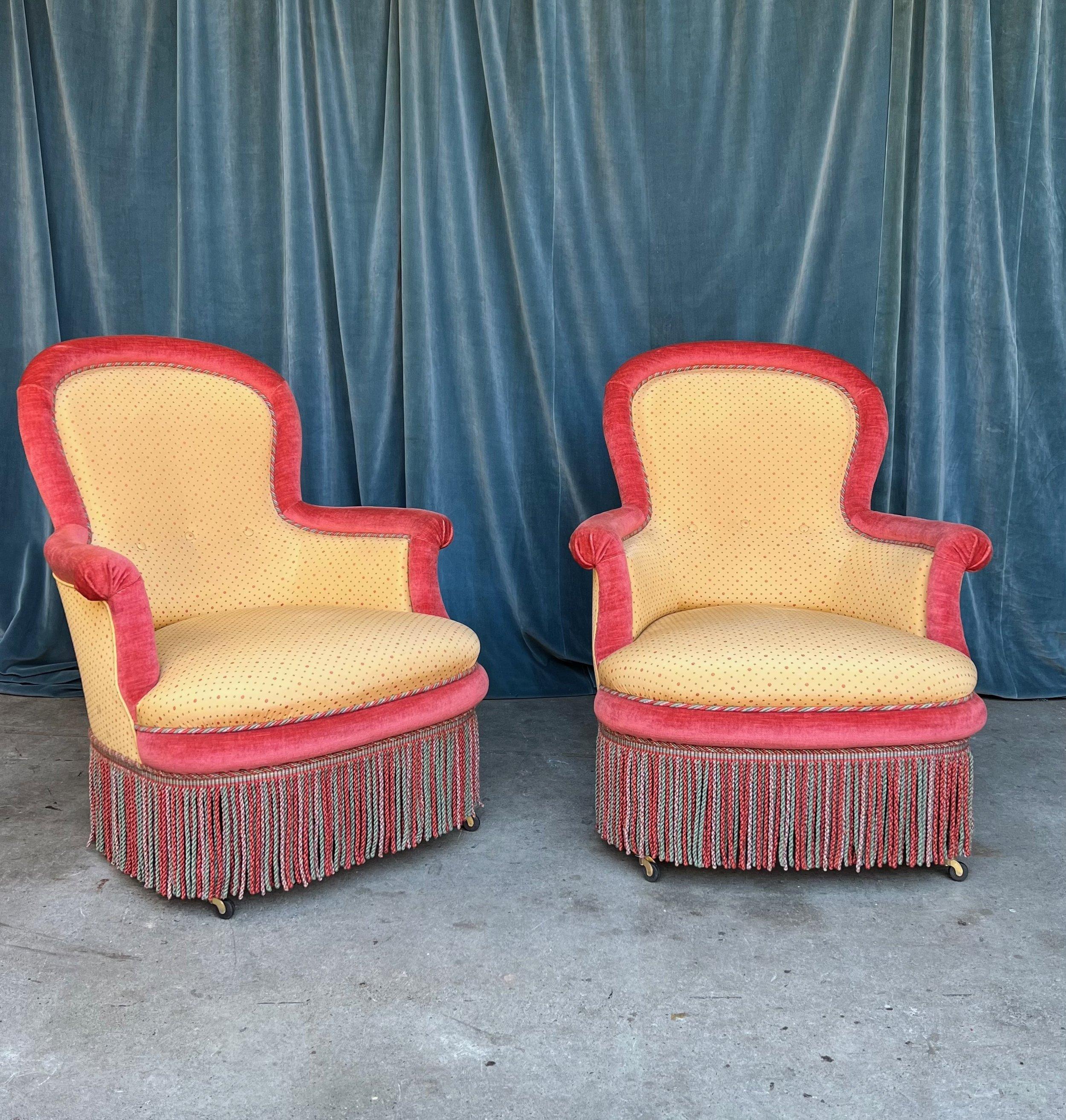 A unique and colorful pair of 19th century French Napoleon III armchairs upholstered in a golden yellow patterned fabric and a contrasting red velvet and featuring multicolored bullion fringe for an opulent final touch. We have added caaters to the