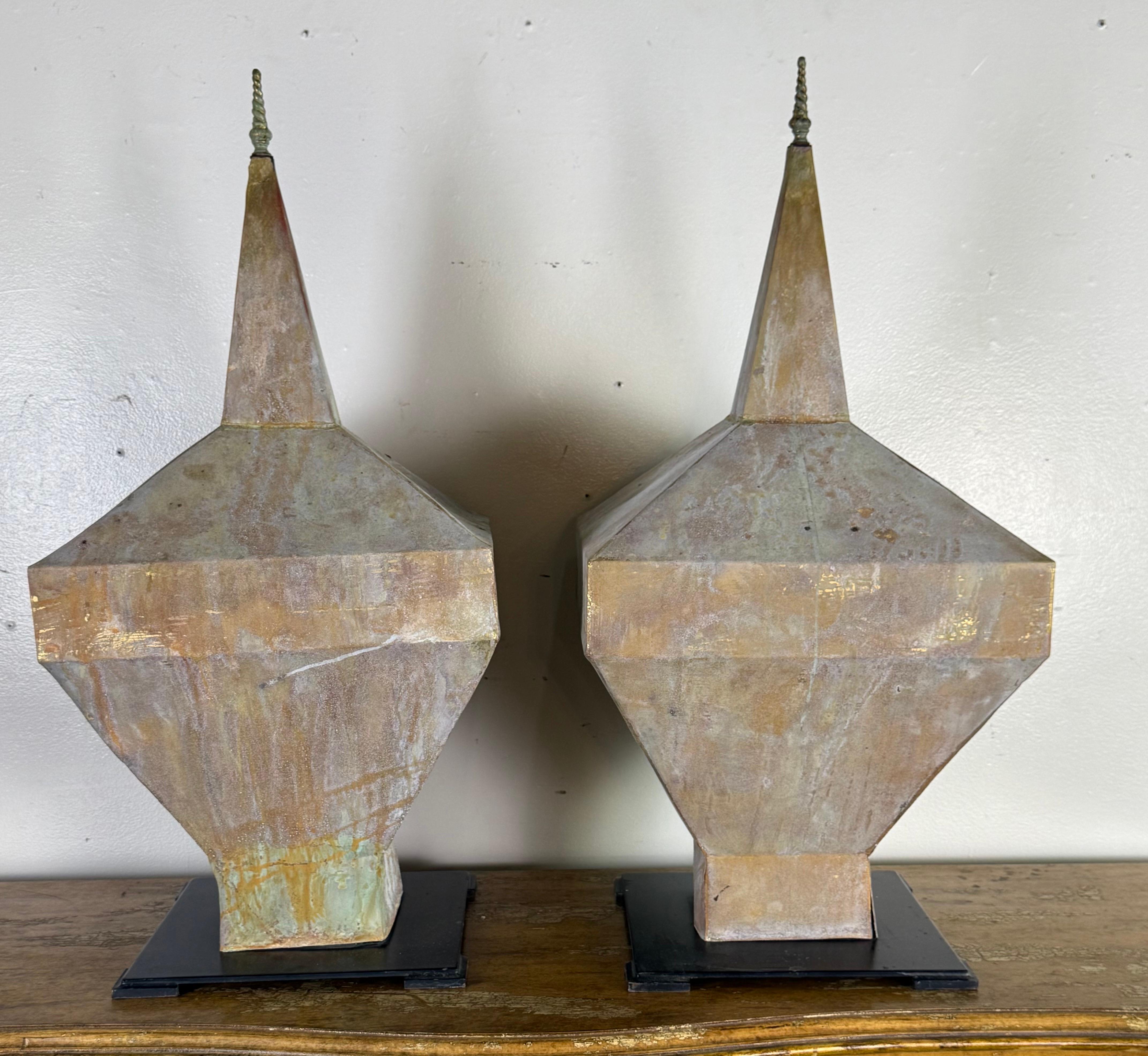 A striking pair of monumental zinc architectural finials, each featuring a tall faceted spire form with a tapered silhouette and twisted metal finial tip. The surfaces display a richly aged natural patina with tones of soft gray, bronze, and