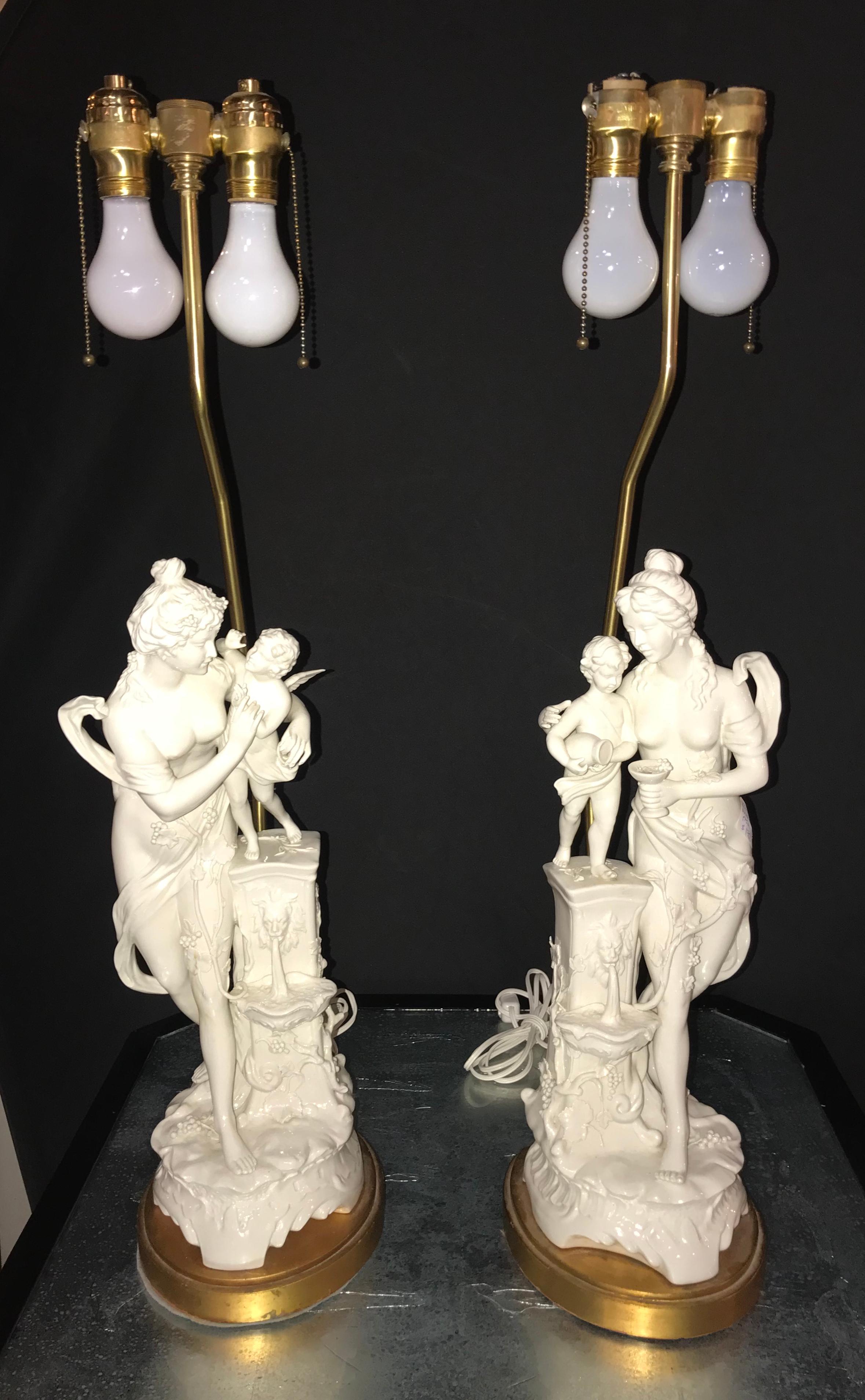 Pair of large porcelain figural opposing woman and child table lamps. This fine pair of antique table lamps are spectacularly done in a white porcelain finish with each bare breasted woman paying attention to a winged angel. One cherub having a wine