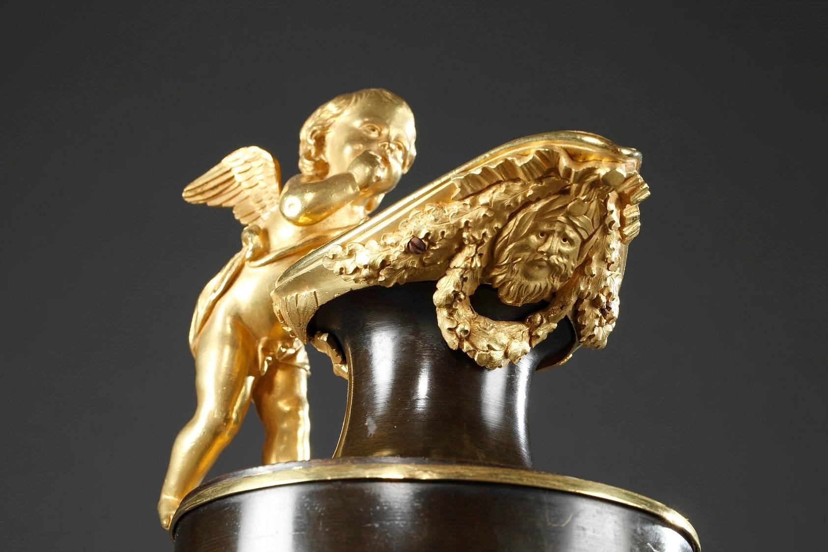 French directory pair of ewers in gilded and patinated bronze with handles in the shape of winged cupids. Both pitchers are decorated with ormolu garlands, a mask, and ribbons on the spout, while the paunch is encircled with a golden band. They rest