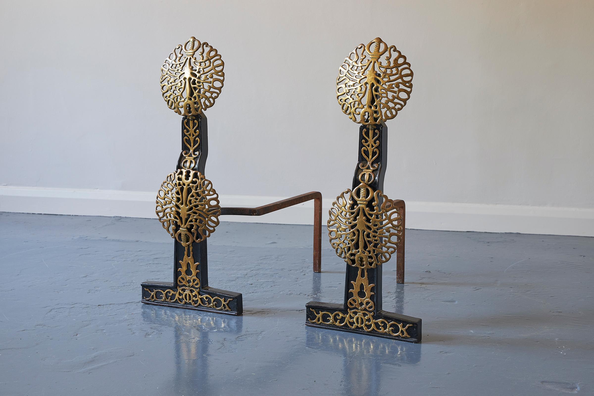 A pair of Art & Crafts brass and iron fire dogs, early 20th century, in the manner of Edward Spencer and the Artificers' Guild. Each with two filigree discs raised on iron supports applied with filigree brass mounts, each with curving billet bar.