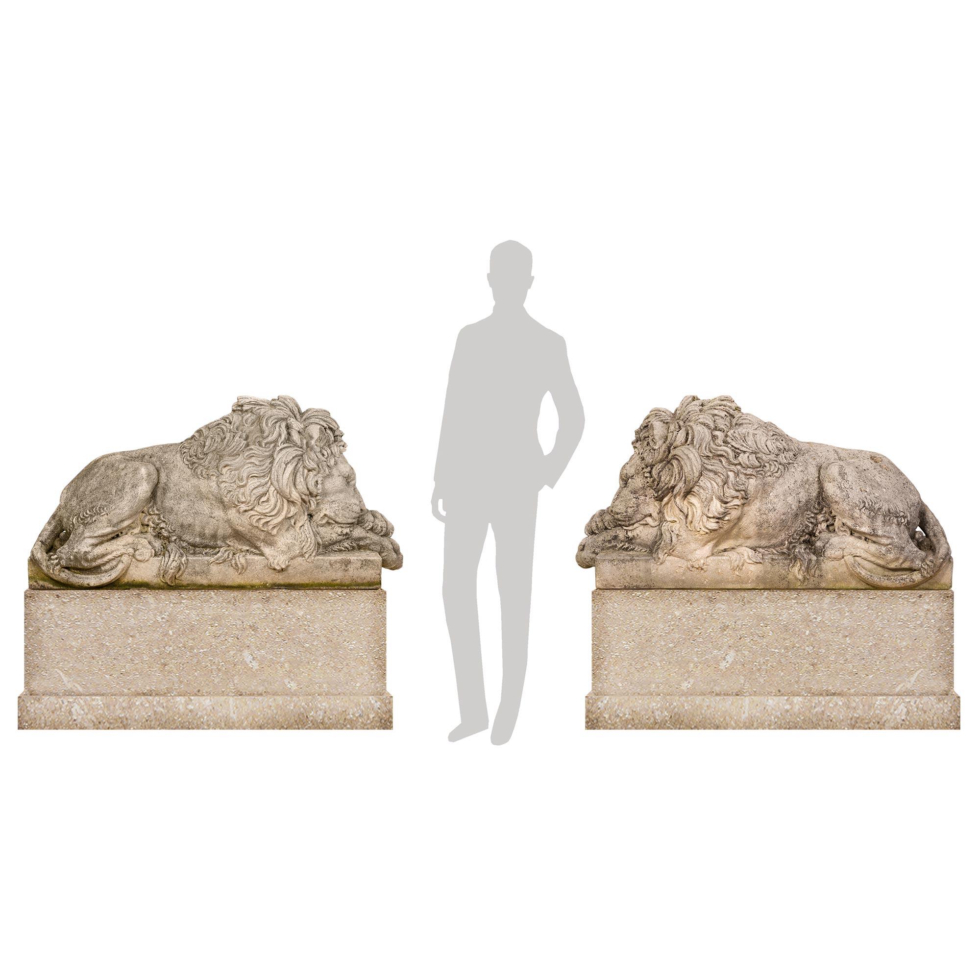 A stunning and most impressive true pair of late 19th century composite stone statues of lions. Each large scale statue is raised by a rectangular base where the handsome wonderfully detailed lions are lying. They each have their paws crossed and