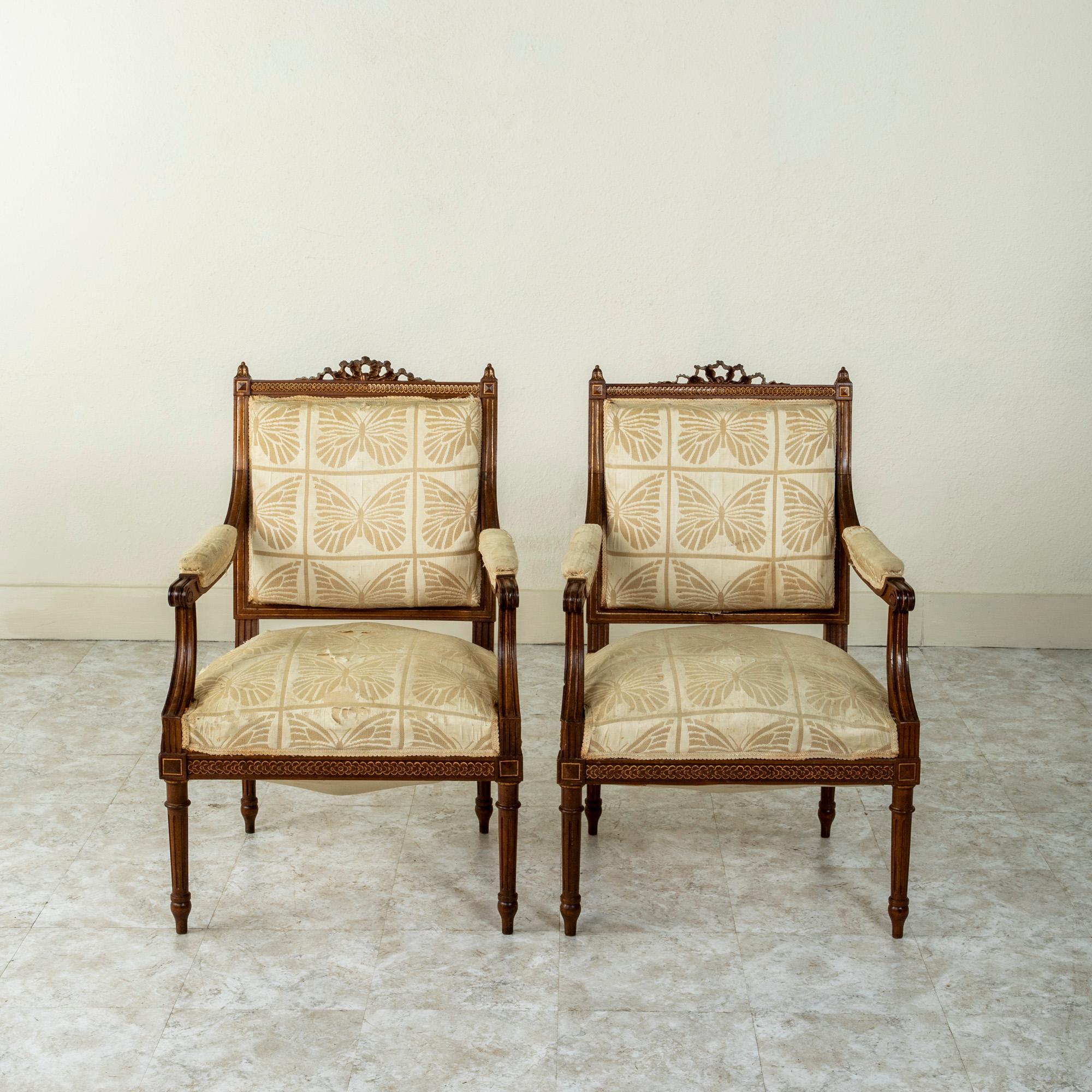 This pair of late nineteenth century Louis XVI style hand carved walnut armchairs features a central knotted ribbon at the top of the seat back and acanthus leaf finials. The chairs are also ornamented with an overlapping quatrefoil motif and gold