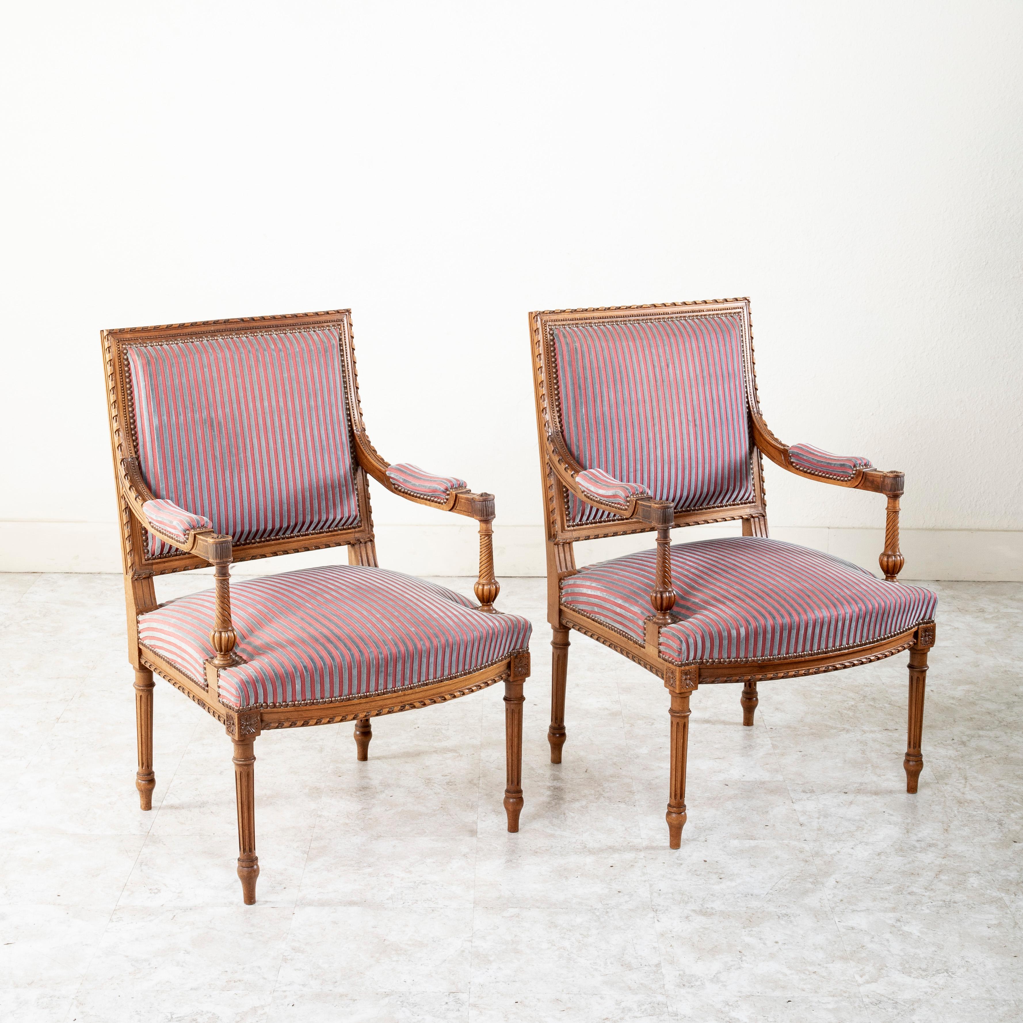 This pair of late nineteenth century French Louis XVI style armchairs is constructed of solid walnut. Their beautifully hand carved details include classic rosettes at the die joints and a twisted ribbon motif around the apron and seat backs. The