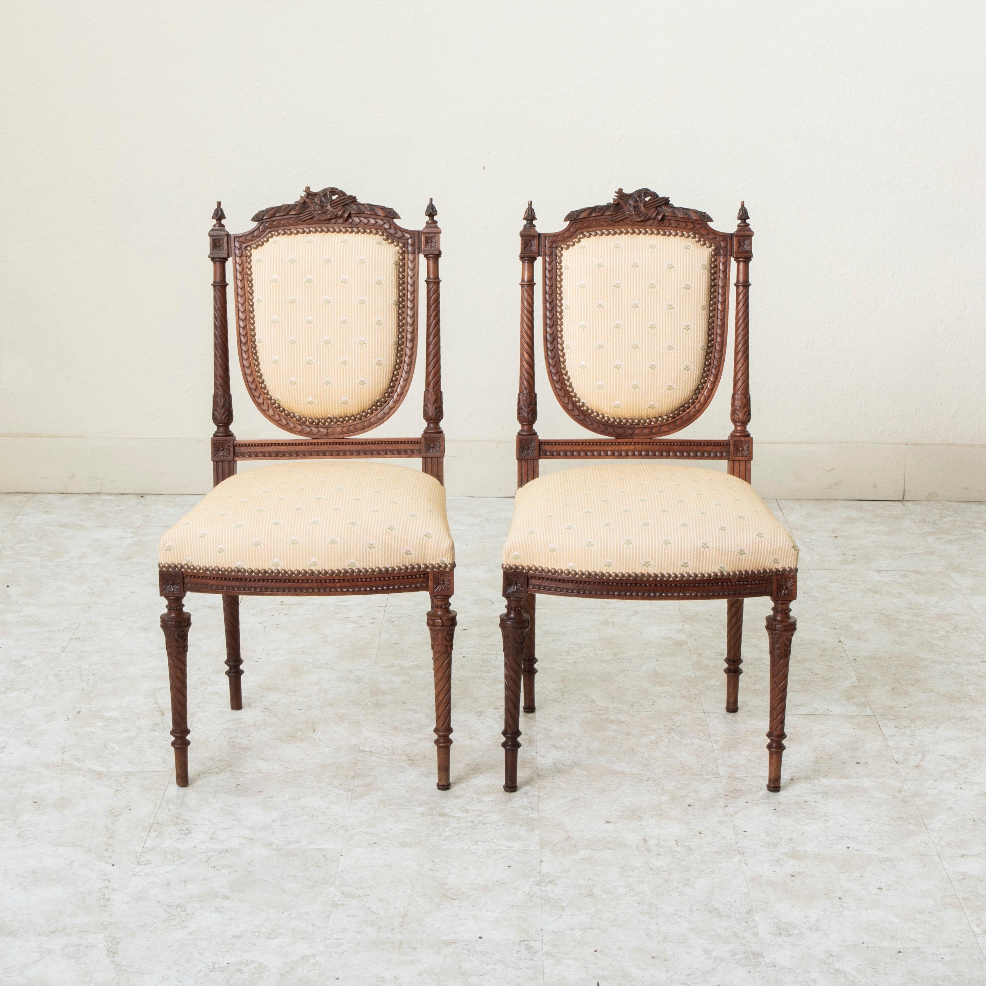 This pair of nineteenth century Louis XVI style walnut chairs features an escutcheon shaped seat back flanked by spiral fluted columns detailed with acanthus leaves. The columns have rosettes at the top and bottom and are appointed with finials. A