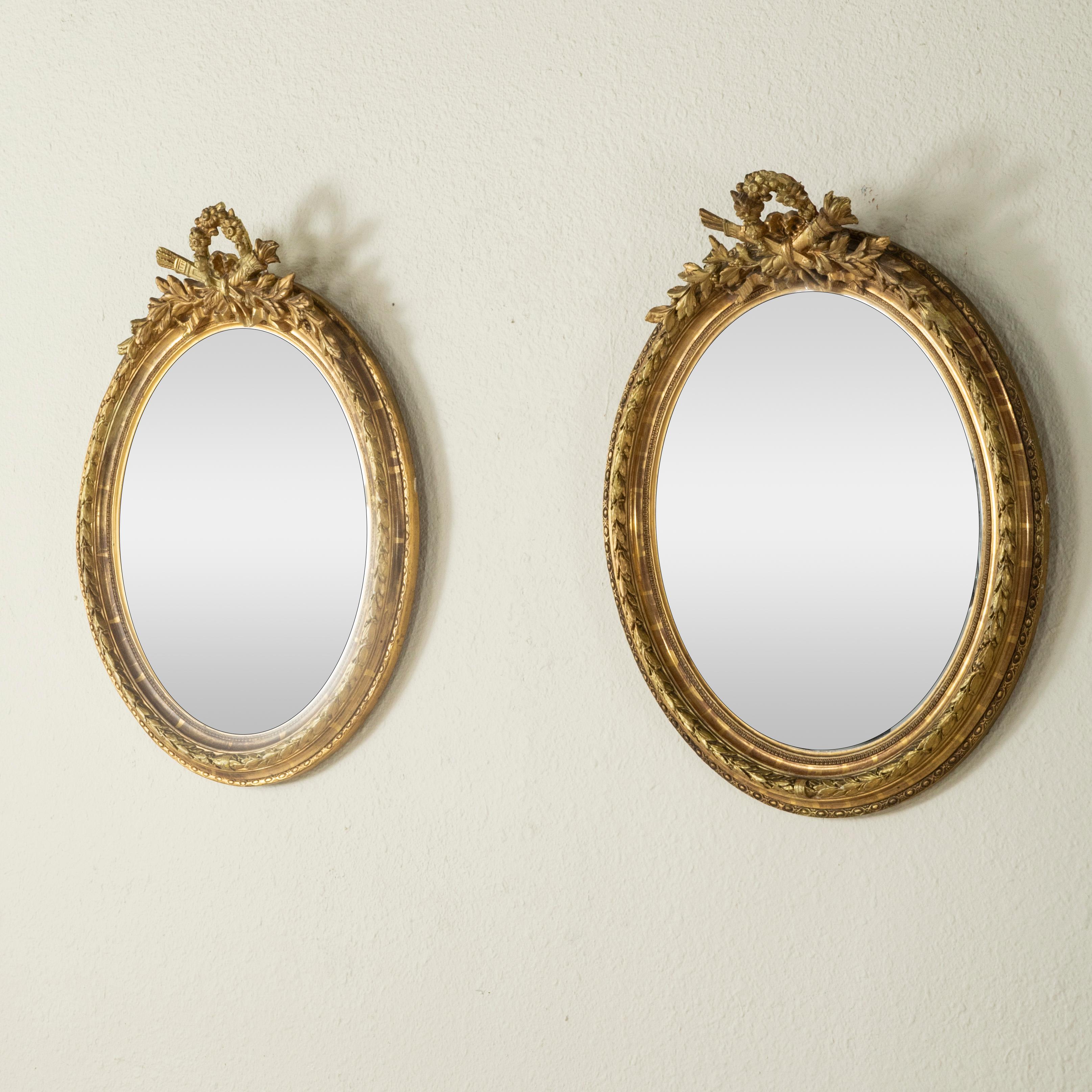 This pair of late nineteenth century small scale oval Louis XVI style gilt wood mirrors features a crown of flowers and a classic crossed torch and quiver at the top. Each frame is detailed with beading, rais-de-coeur, and a laurel pattern. The
