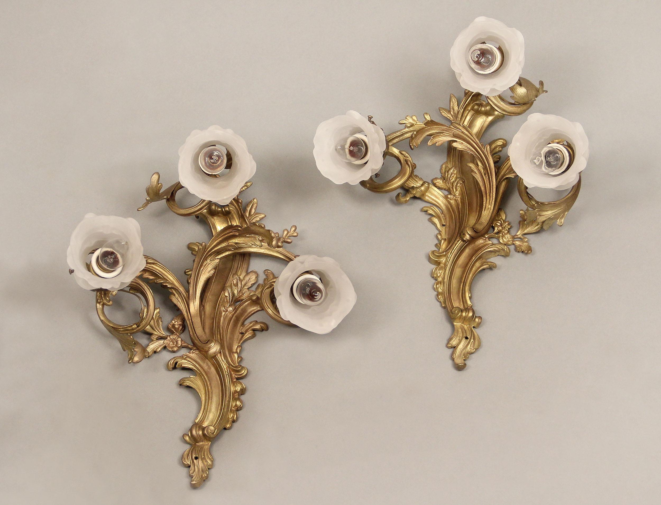 A pair of late 19th century gilt bronze three-light sconces

Rococo style sconces with the backplate and arms decorated with flowers and foliage, the lights with rose shades.