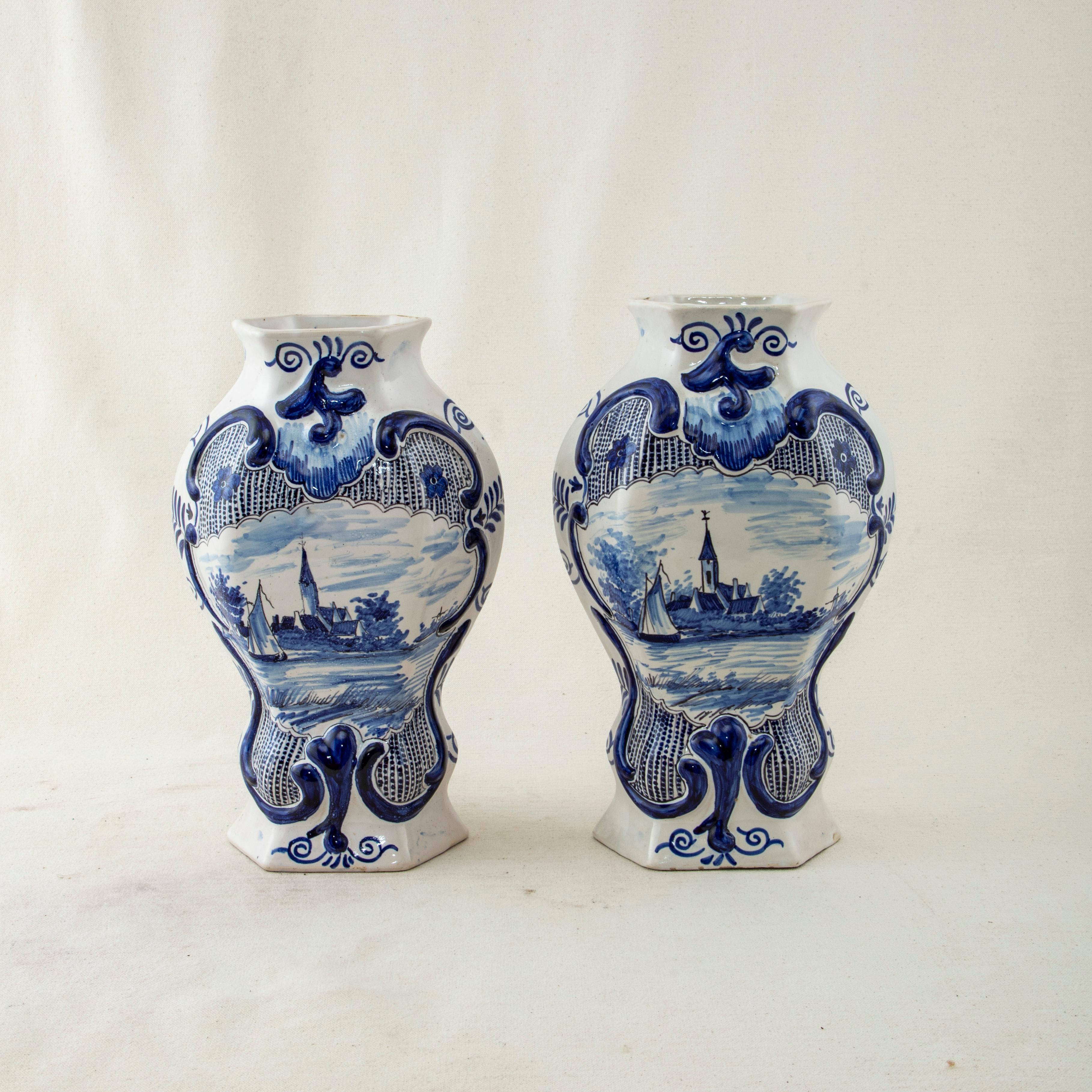 This pair of late nineteenth century blue and white delft vases features hand painted scrolling and a central scene of a boat on a river with a small church in the background. On the backs of the vases is a stylized leaf design. The bottoms are