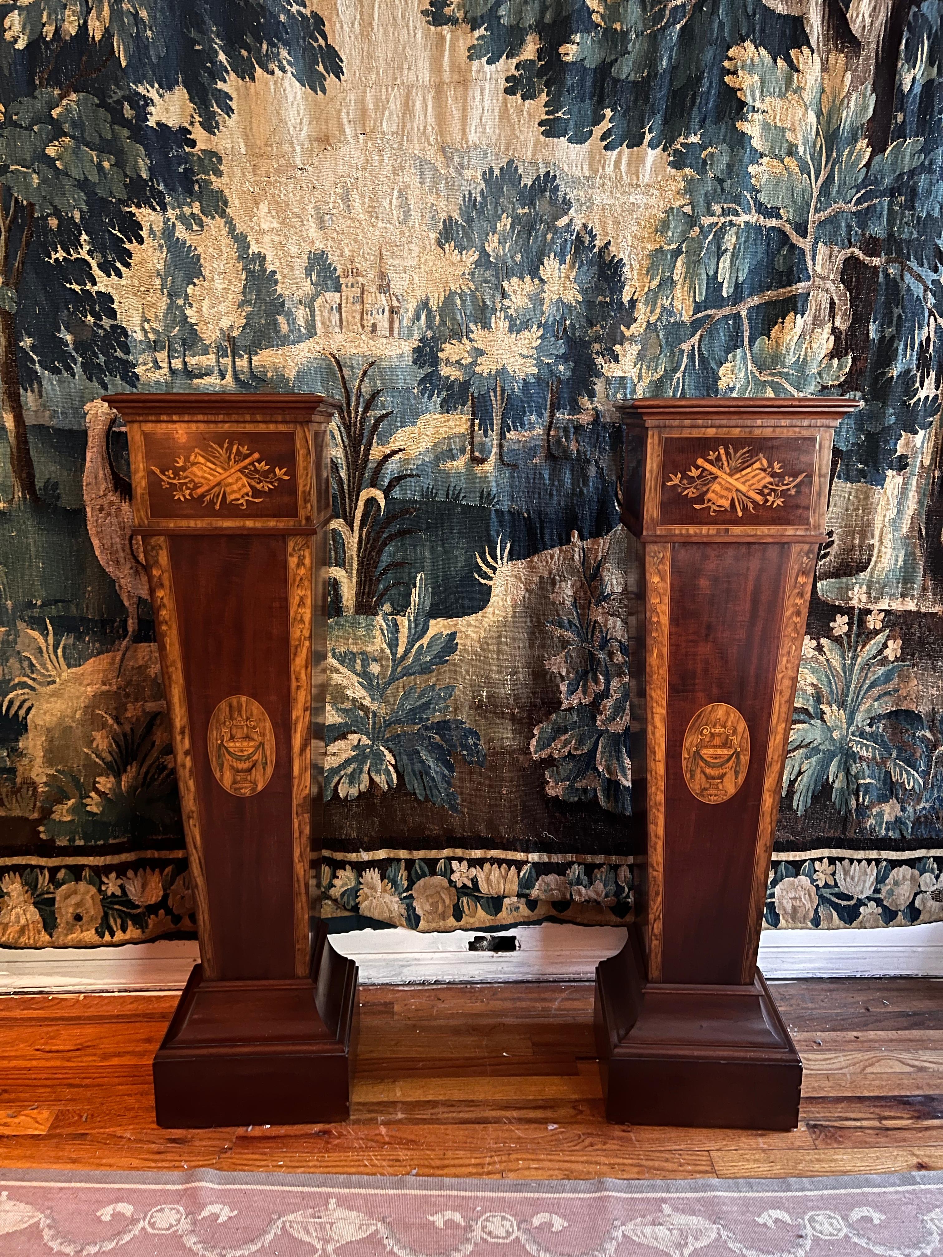 A pair of handsome neoclassical pedestals in mahogany and inlaid detailing. The upper portion is decorated with musical instruments and foliage with the shaft bearing a oval inlay of a classical urn. The edges of the shaft are decorated in with