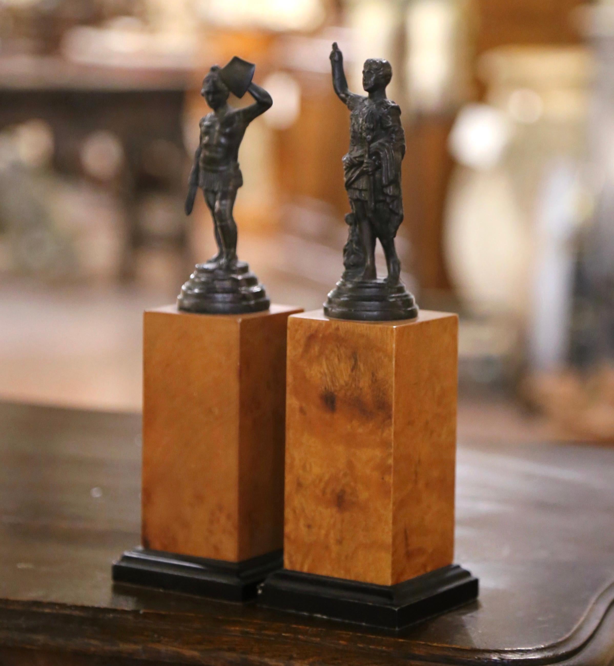 Accessorize an office or library shelving with this pair of small bronze statues mounted on wooden plinths. Each statue depicts a figure from classical antiquity: the one on the left appears to be a Roman gladiator, holding a shield and wearing a