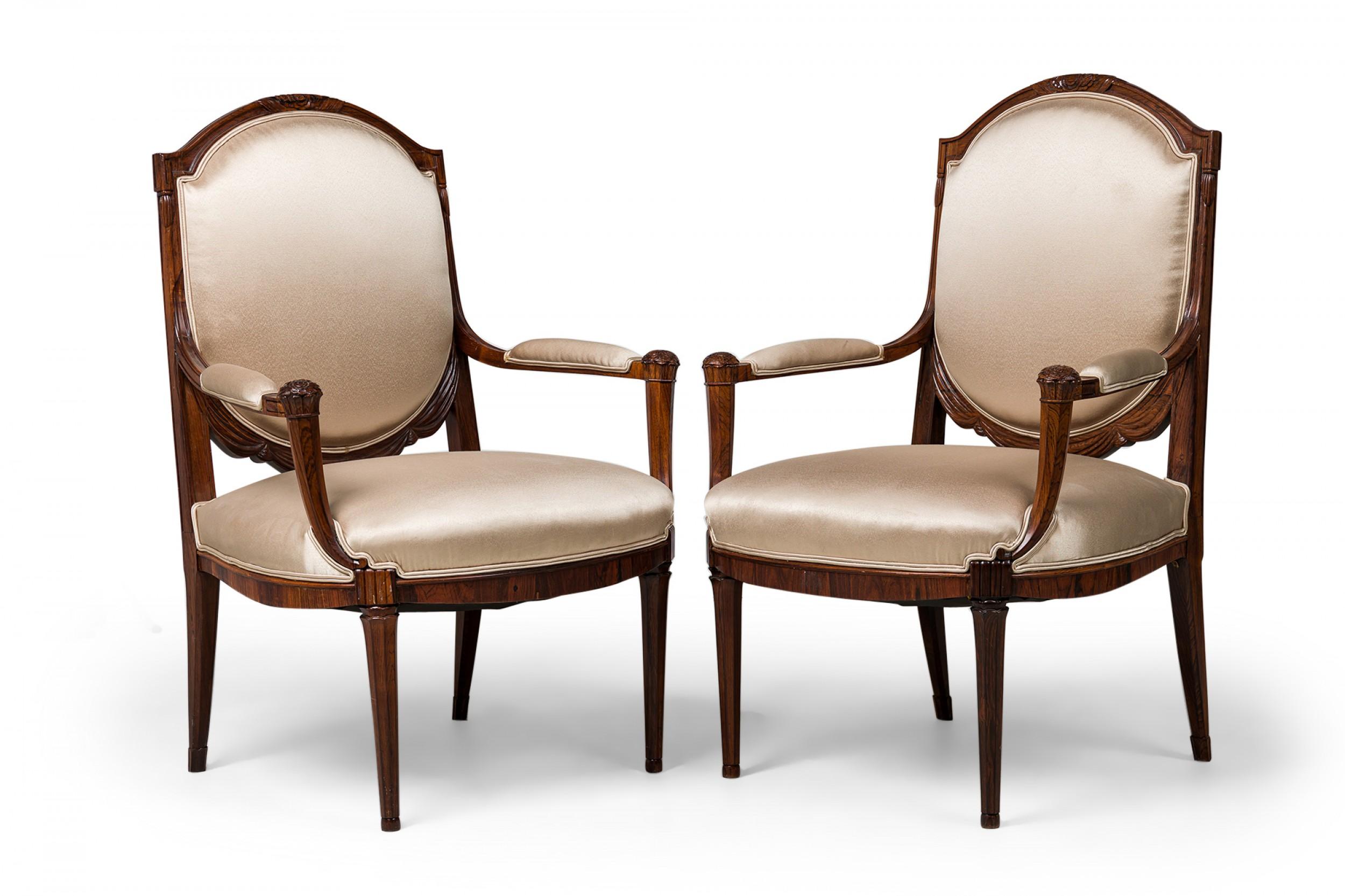 Pair of Late Art Deco French mahogany armchairs featuring an oval back in a carved arch foliate design with drapery and rosettes at the arms\' corners, upholstered in a beige sateen fabric with double piping, standing on 4 molded legs, the back two