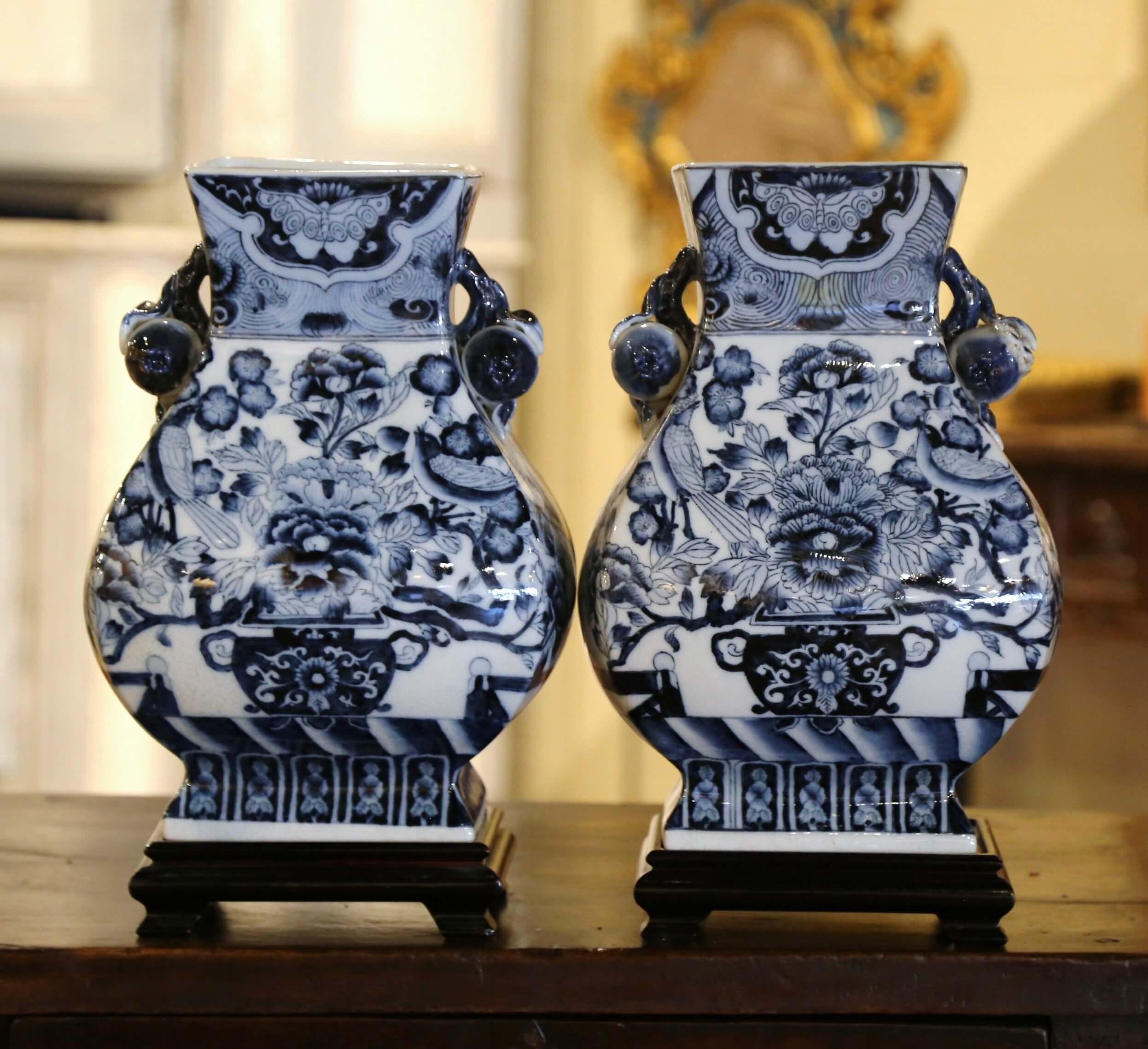Decorate a mantel or a console with this elegant pair of antique vases. Crafted in China circa 1980, each porcelain urn-shaped sits on a dark wooden base and features an octagonal shape and a substantial presence. The large vase is decorated with