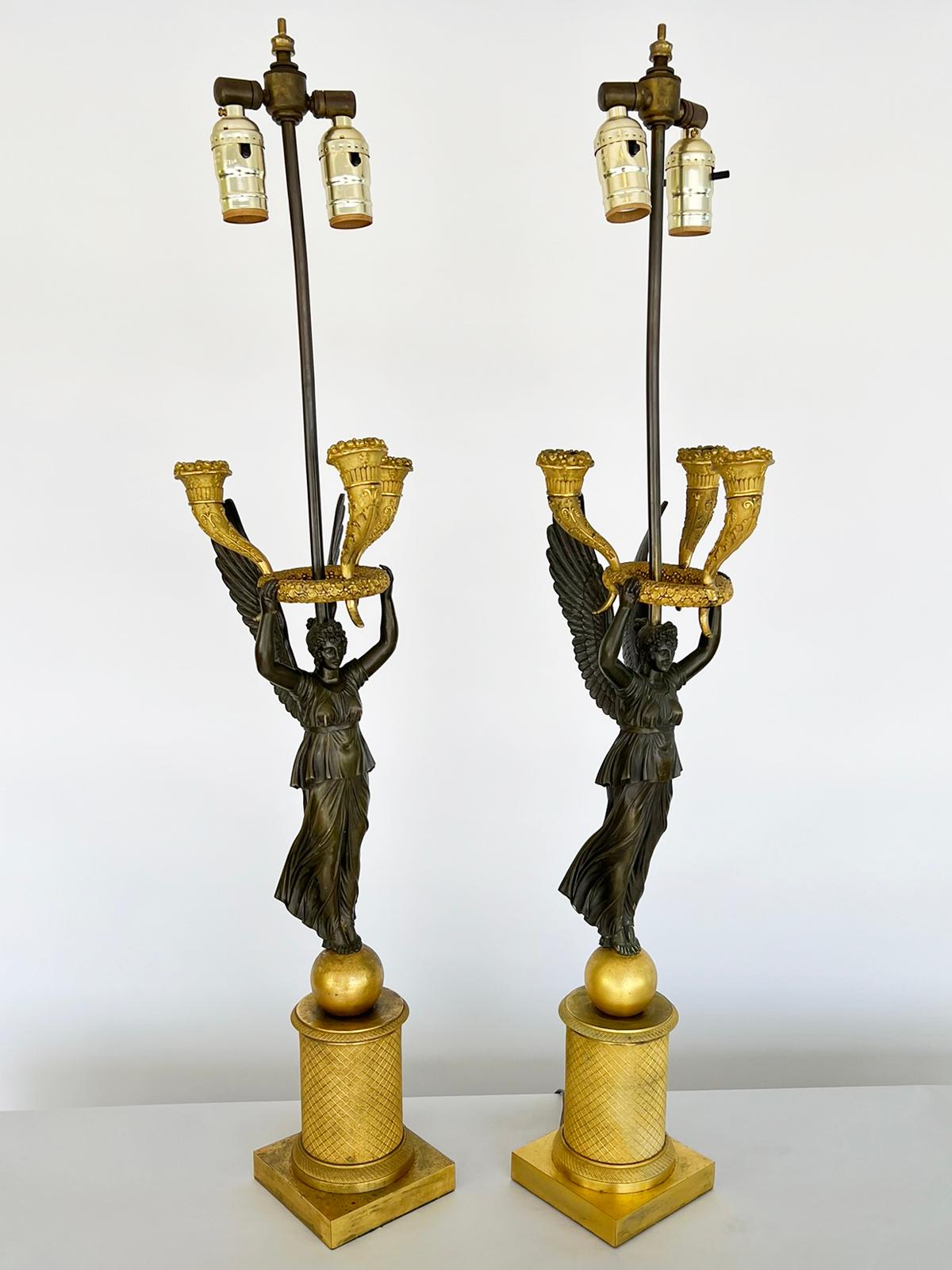 Pair of period Empire figural candelabrum. Each modeled as a winged figure of Victory, of patented bronze, with both arms supporting a floral wreath, and three horns-of-plenty, of ormolu. Each caryatid stands upon a sphere atop a square plinth,