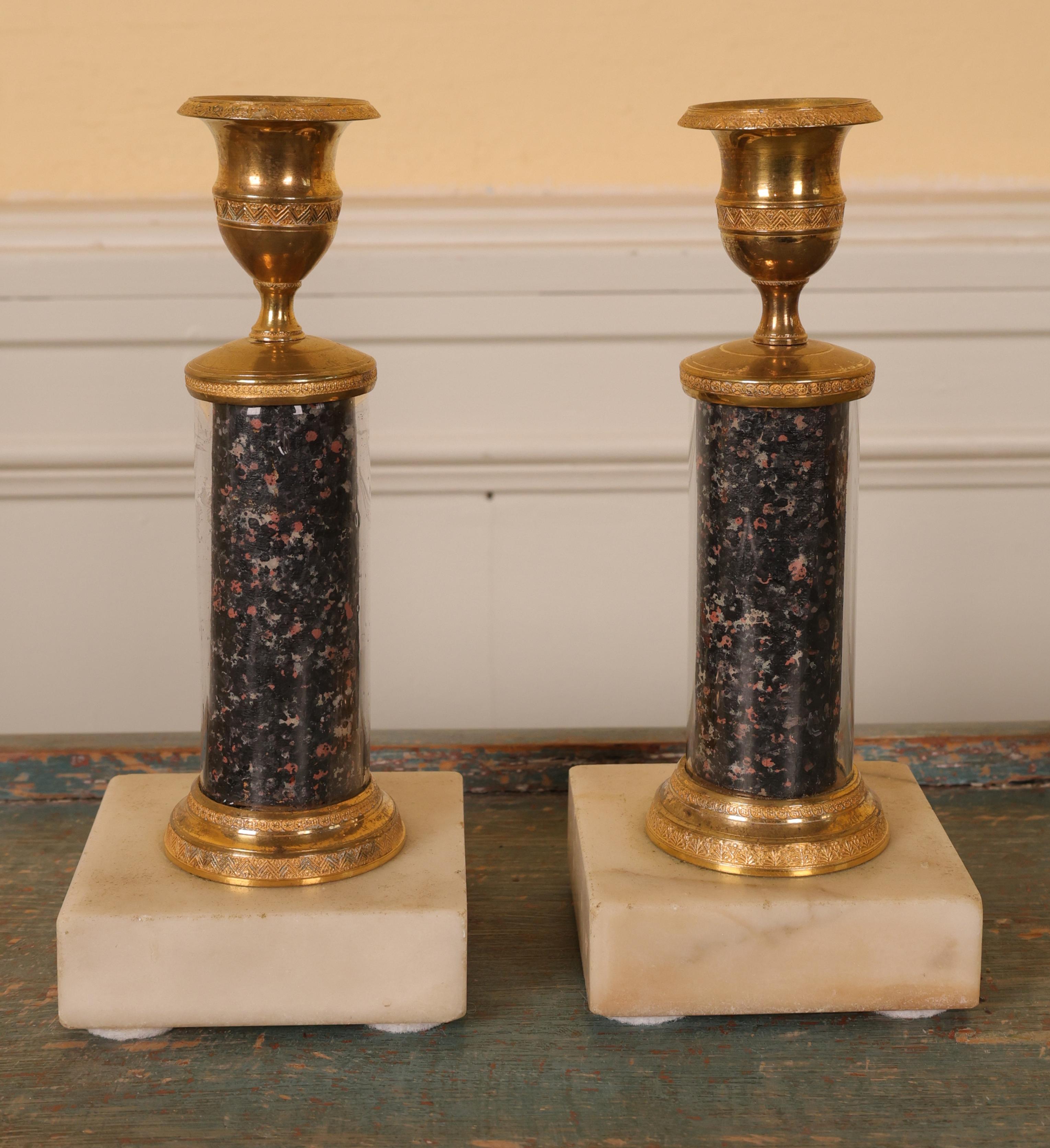 A pair of late-Gustavian candlesticks made in Stockholm around 1800. The stems are made of glass columns with a porphyry-imitation paper on the inside. The fittings and light sockets are burnished gilded bronze. The bases are made of white Carrara
