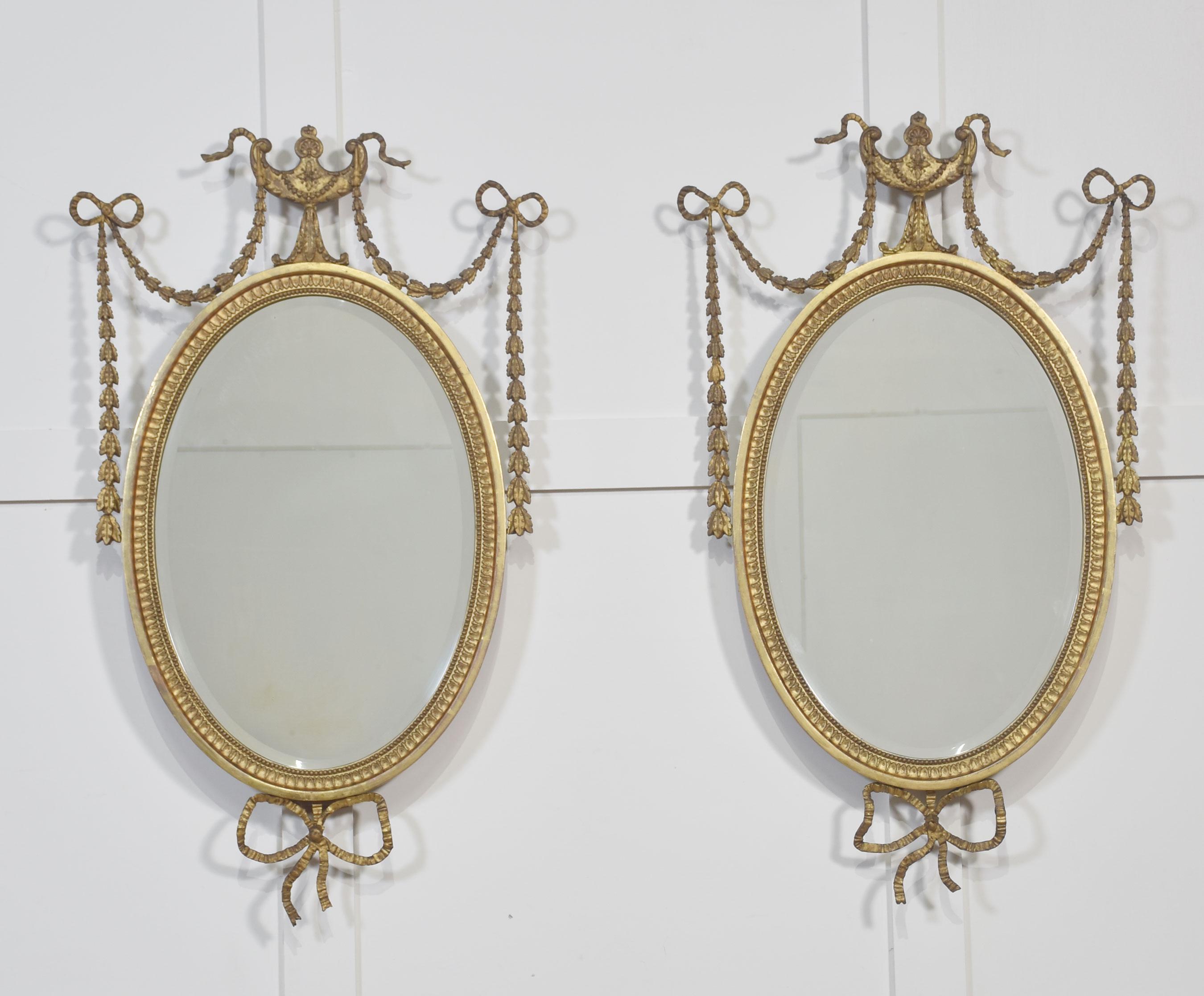 An exceptional pair of late Regency giltwood oval wall mirrors, circa early 19th century. Each mirror retains its original beveled glass, set within a finely carved gilt frame with beaded and foliate detailing. The crest features a classical urn