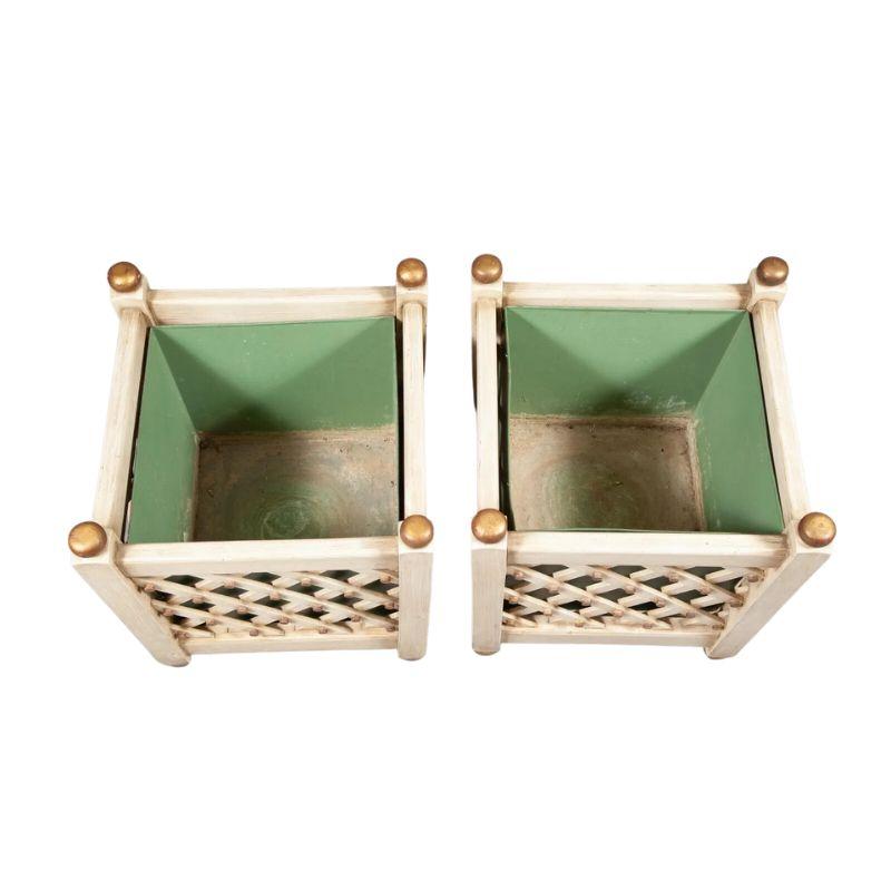 A pair of painted wood square planters with a diagonal lattice motif on each side revealing a green plant insert to hold a topiary, plant or greenery.  The planters are painted white with gold accents to the round finials at each corner, round gold
