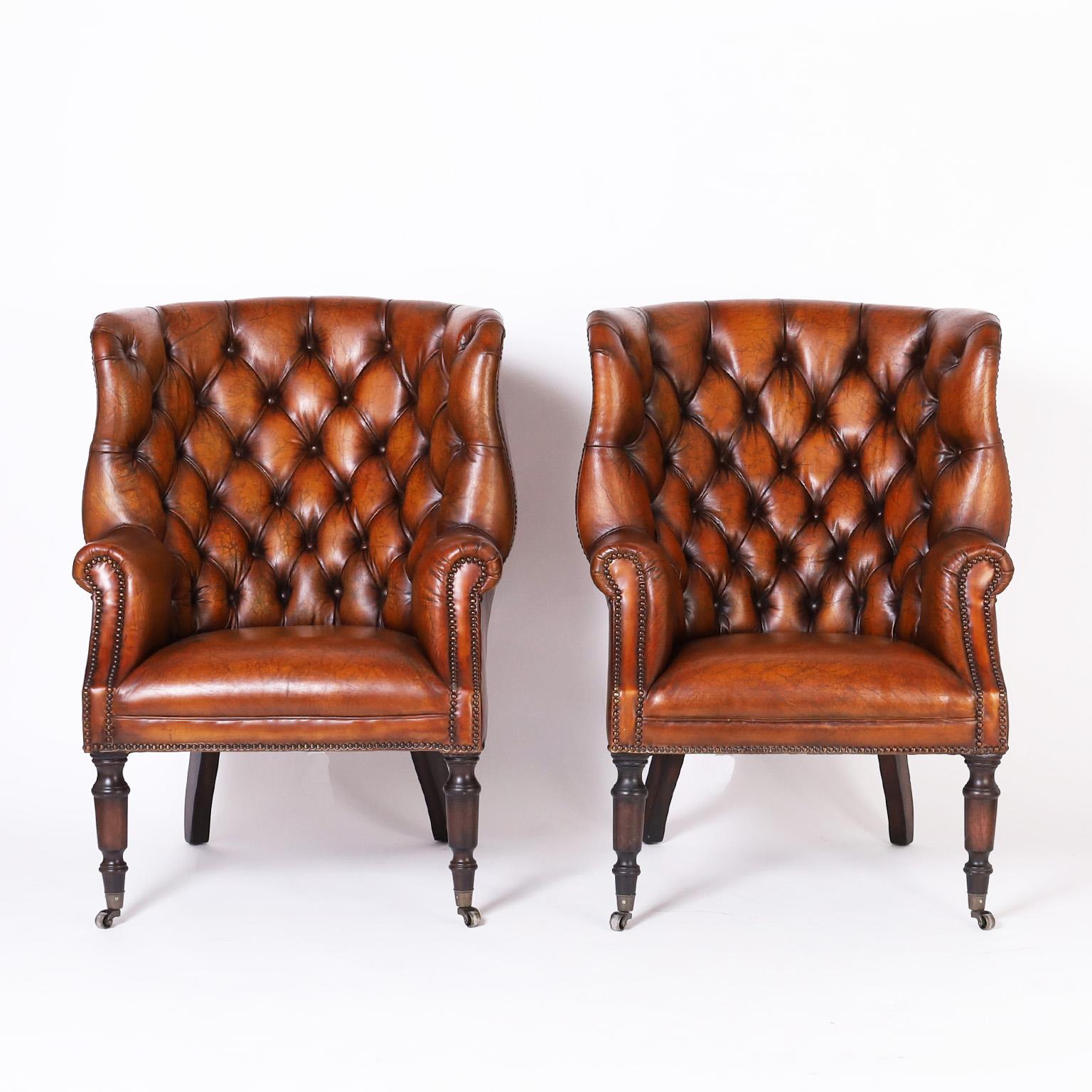 Handsome pair of British colonial style wingback armchairs upholstered in lush brown leather in a classic elegant form and featuring button tufted backs, brass studs, and turned legs on metal casters.