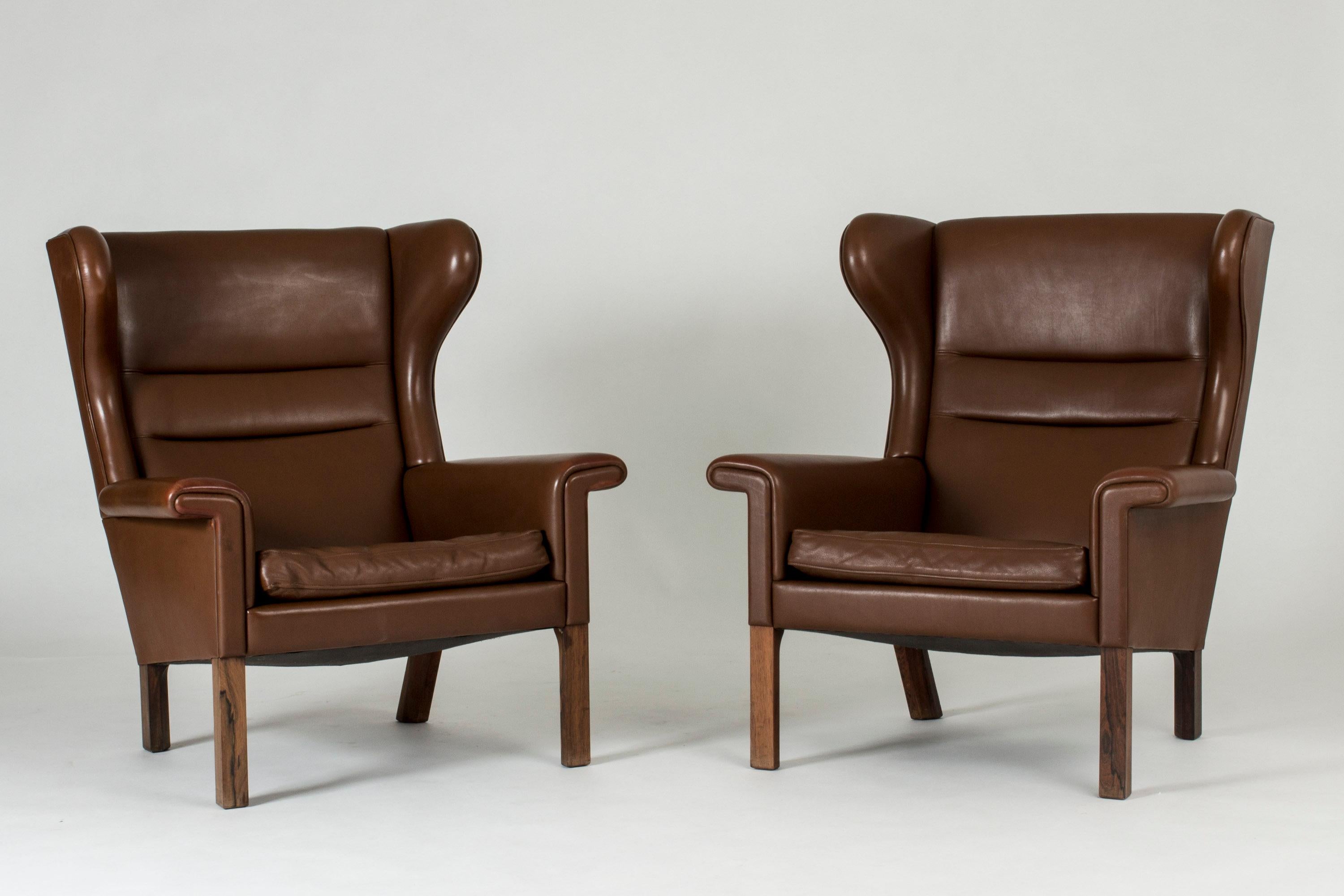 Pair of elegant wingback lounge chairs by Hans J. Wegner, made with dark brown leather and rosewood legs. Pair of ottomans made from rosewood with loose leather cushions. The original tags, including the name of the consignee “Arkitekt Sv. Aa.