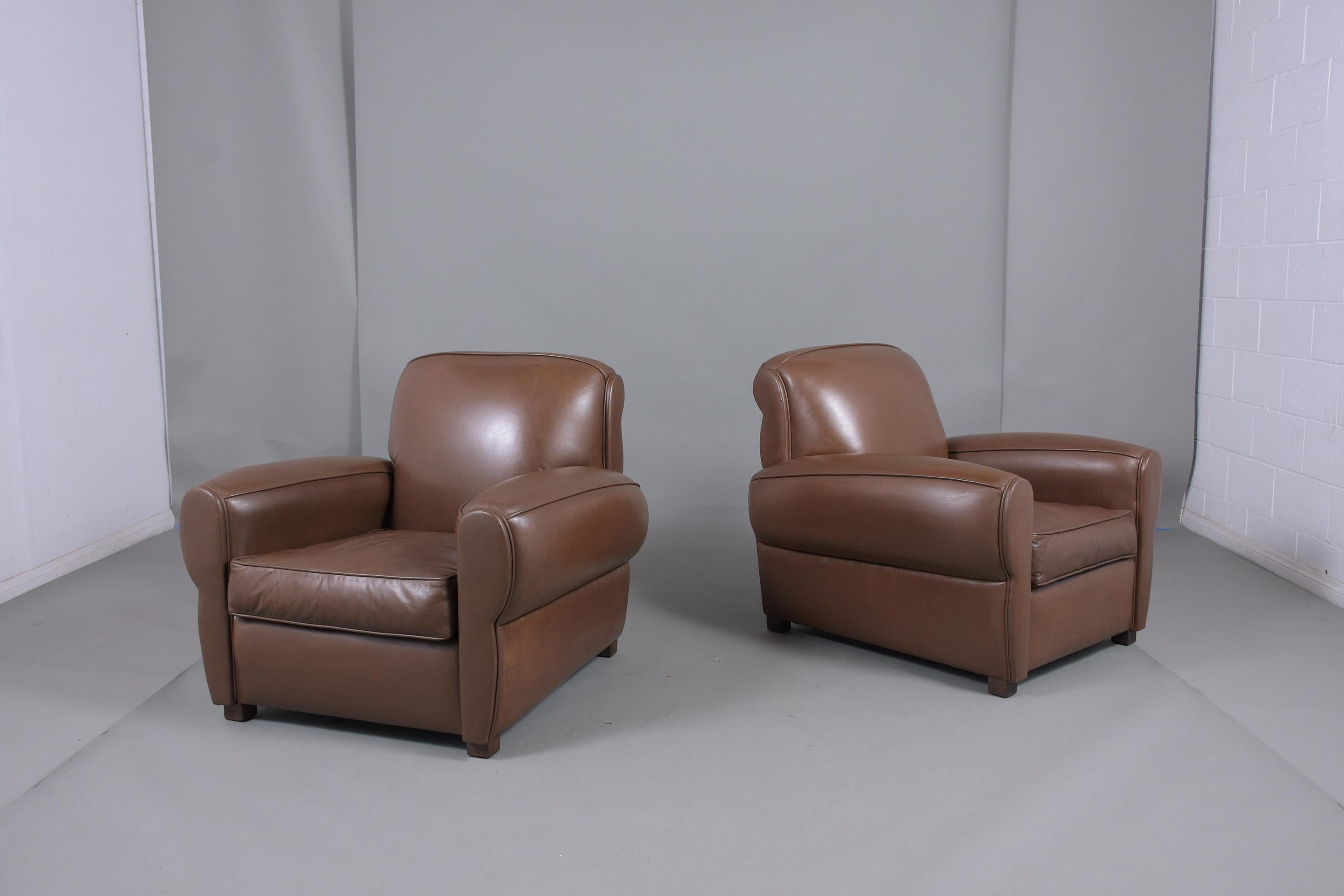 An extraordinary pair of vintage Art Deco-style club chairs are crafted out of wood and leather and restored by our team of expert craftsmen. This fabulous set of lounge chairs features the original brown leather upholstery and is in a good