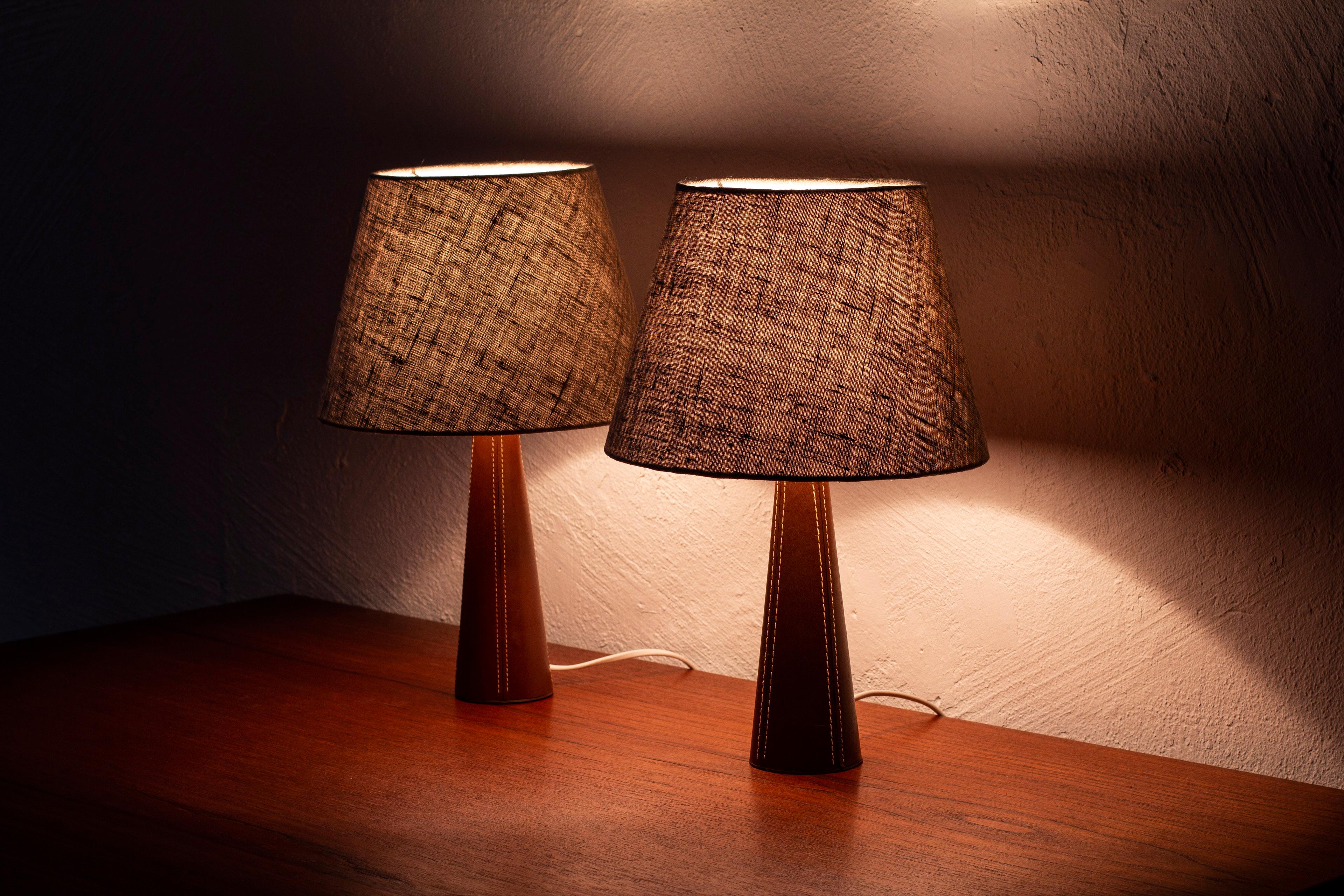 Pair of leather clad table lamps similar to the style of Lisa Johansson Pape. Conical lamp base with brown hand sewn leather finished with a brass lamp holder. Lamp shades in natural/grey linen fabric. Made in Scandinavia during the 1960s.Light