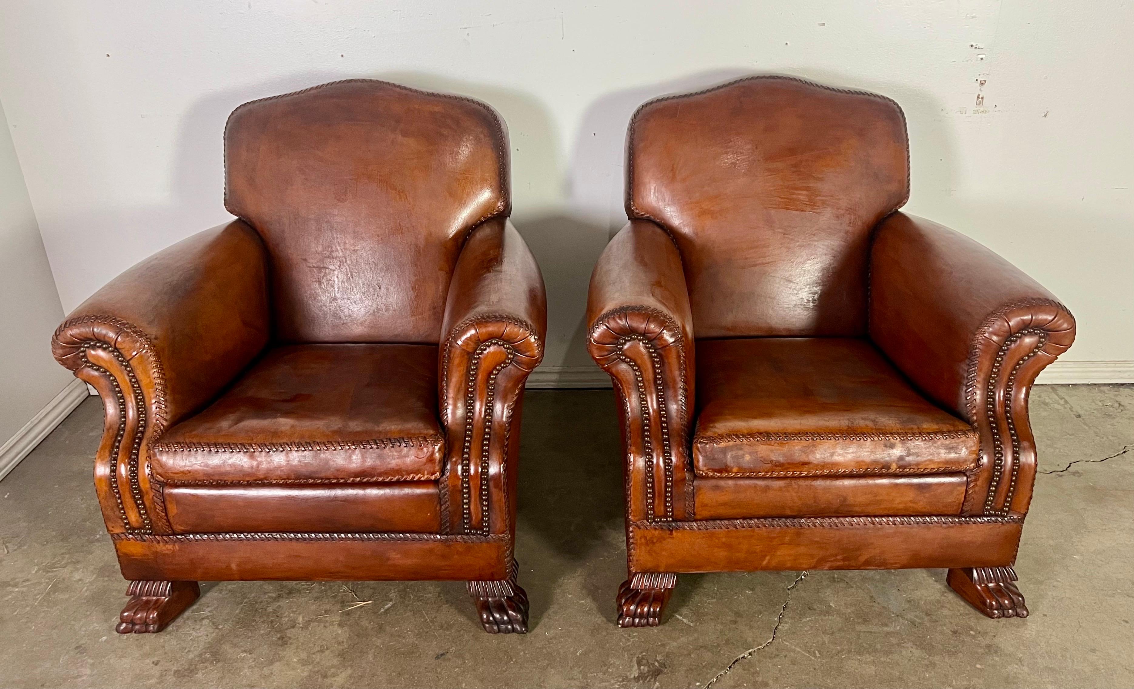 Pair of classic leather club chairs with a vintage aesthetic.  They feature a rich brown leather upholstery with a well-aged patina, giving them a timeless and sophisticated appearance.  The chairs have rolled arms with intricate nailhead trim