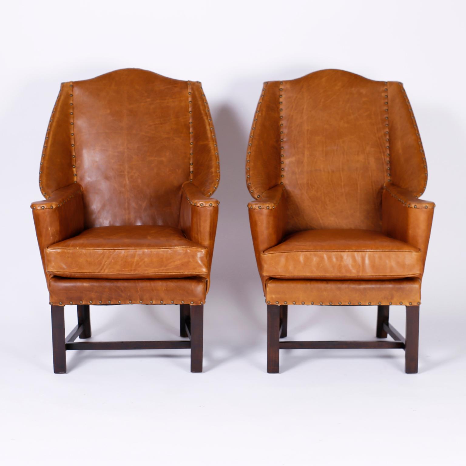 Pair of British colonial wingback chairs upholstered in soft comfortable tan leather highlighted with brass studs and set on Classic Chippendale style legs.