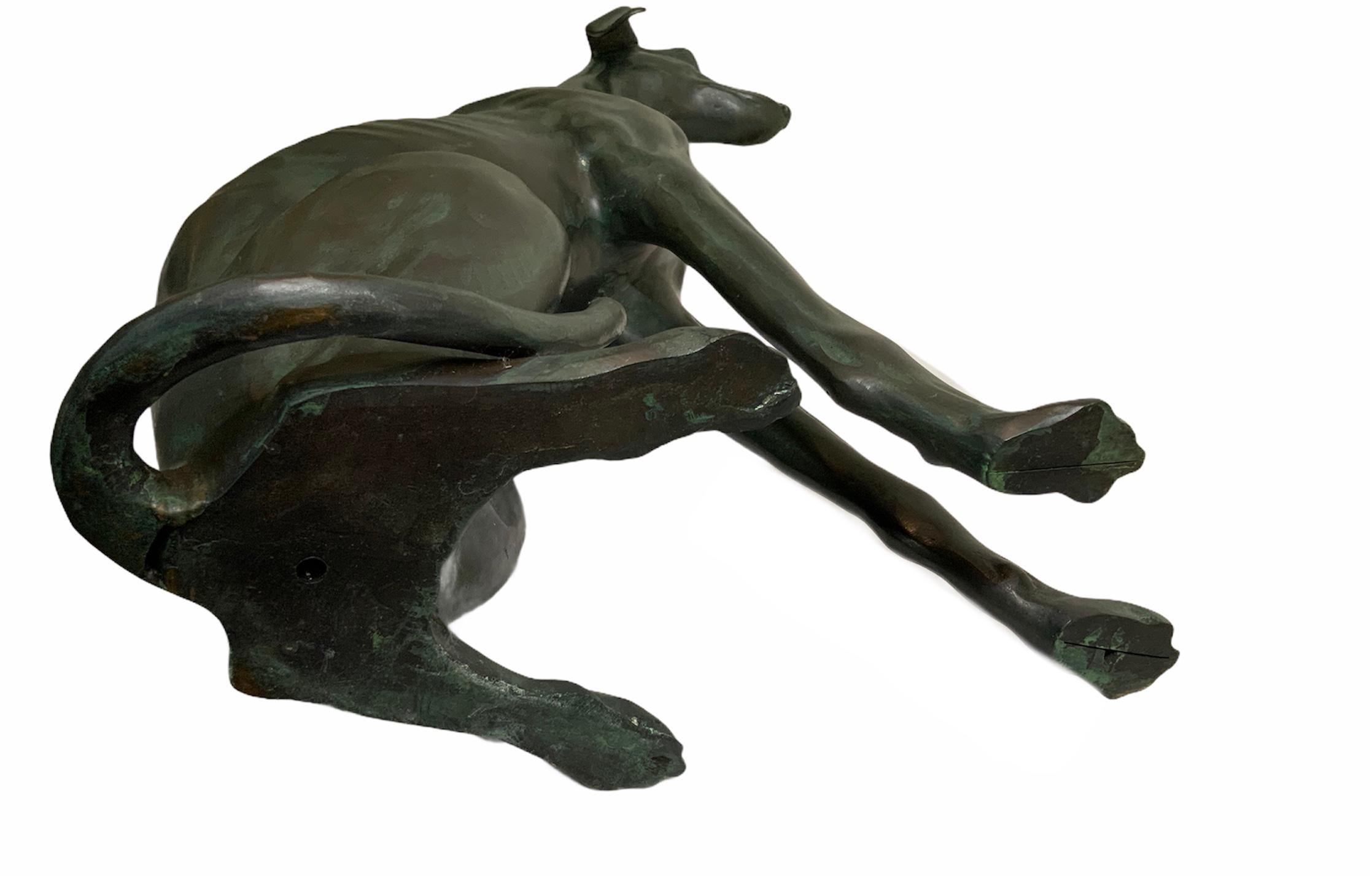 This is a pair of patinated bronze whippets dogs. One of the dog is sitting down and its right ear is lifted up. The other one is very relaxed with its long frontal legs stretched. They are very well done and every detail of its body appears real