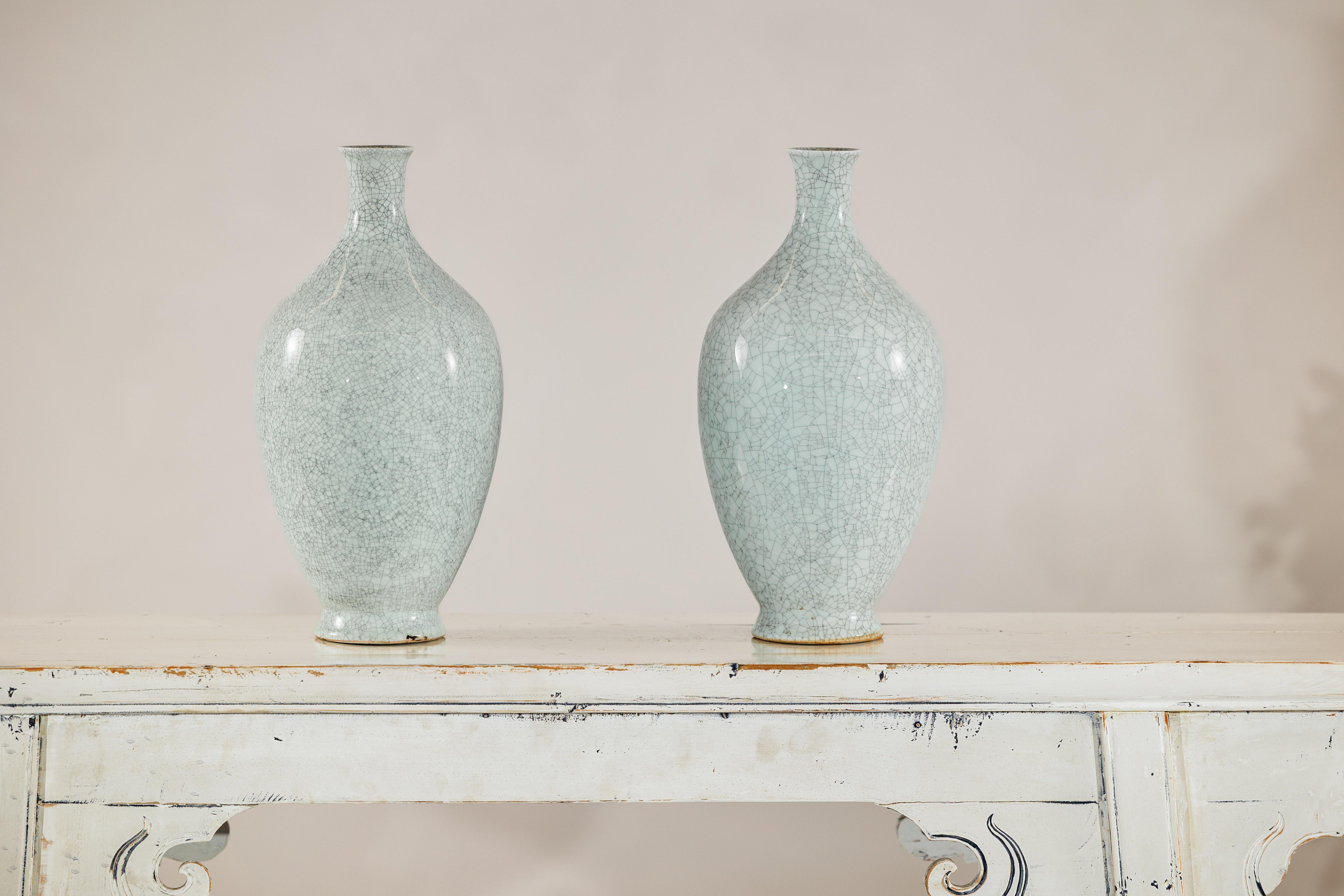 This elegant pair of porcelain vases features a light celadon grey glaze enriched by a dense and intricate crackle pattern that gives each form a refined, timeworn texture. Standing at 14.5 inches high with a 7-inch body diameter and a delicate