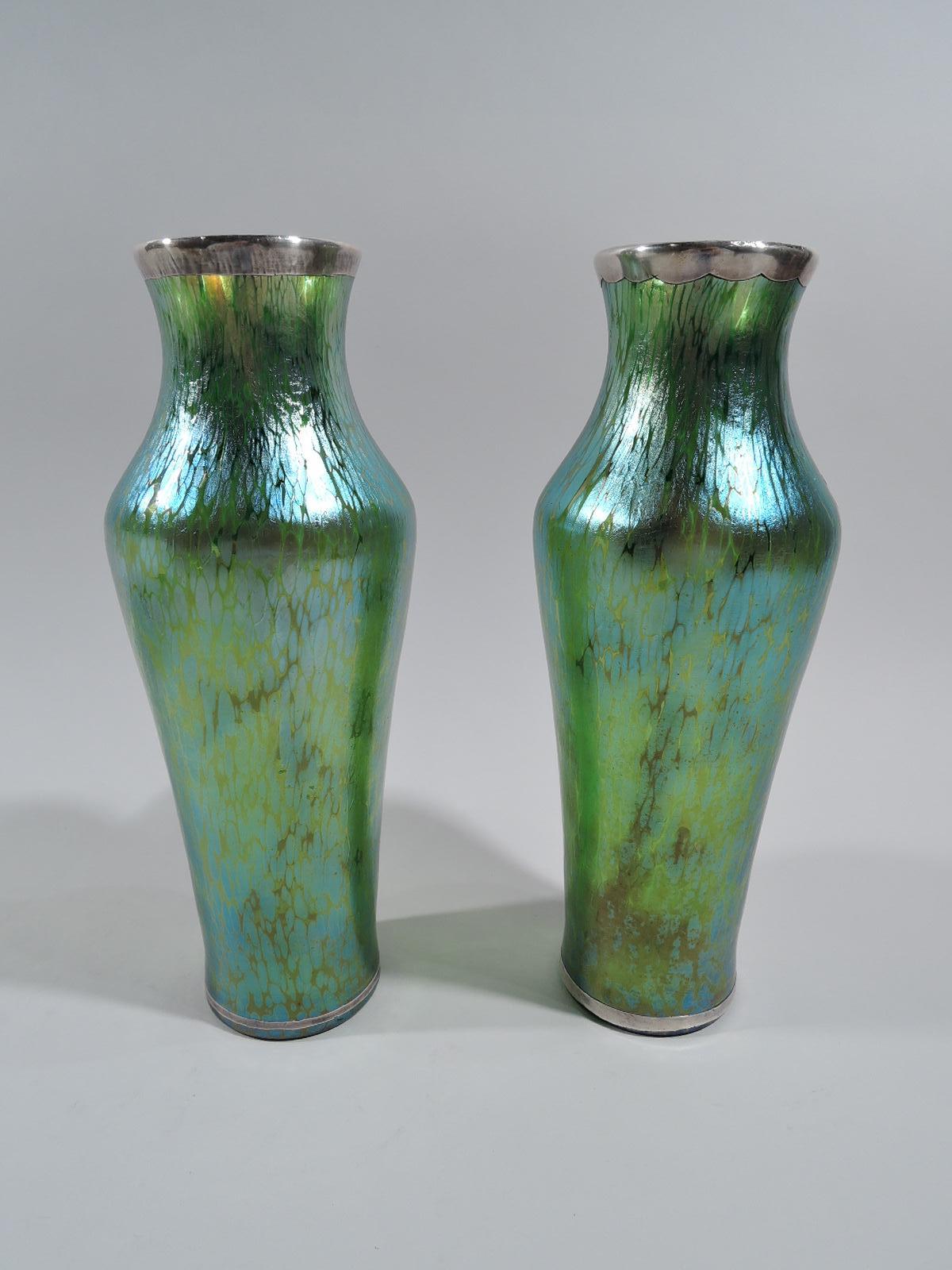 Pair of turn-of-the-century art glass vases by historic maker Loetz. Each: Slender with tapering sides and inset neck with flared mouth. Glass iridescent green with oil-style fracture pattern. On front is engraved silver overlay in form of