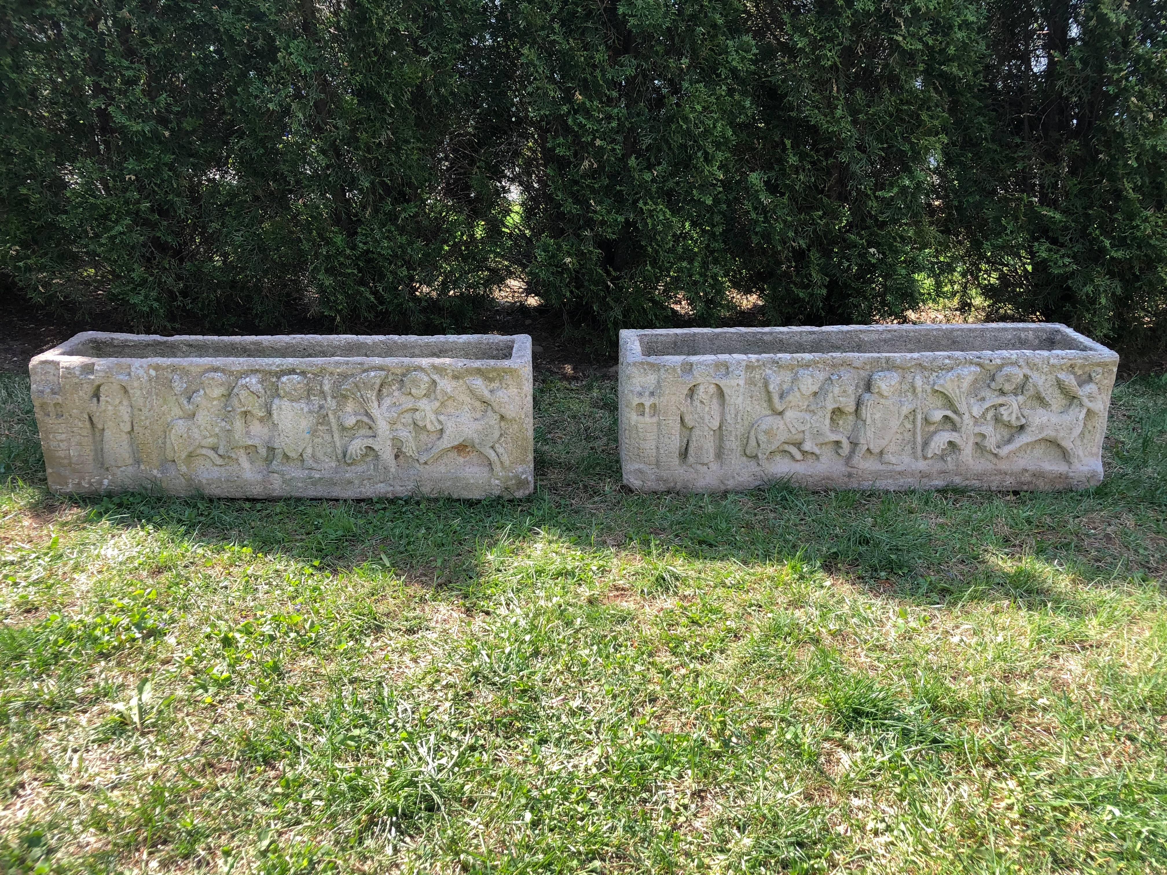 This long pair of cast stone jardinières features a creamy white surface with light gray weathering, and an intricate panel of deeply cast bas relief figures on the front side that tell a story. From the left-hand side, we see the outline of a