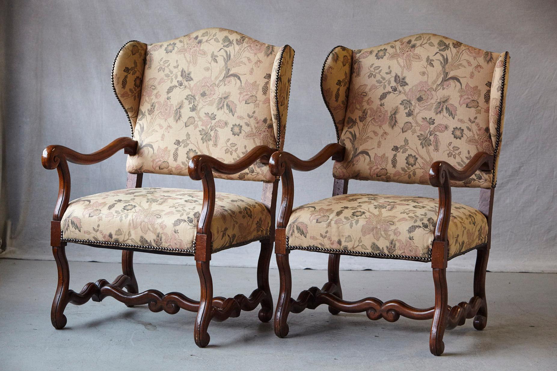 Striking pair of antique French Louis XIV style, walnut wingback fauteuils with carved armrests, standing on 'os de mouton' legs joined by H-shaped stretcher base, upholstered with a tapestry like fabric with nailhead trim, nailhead trim.
These