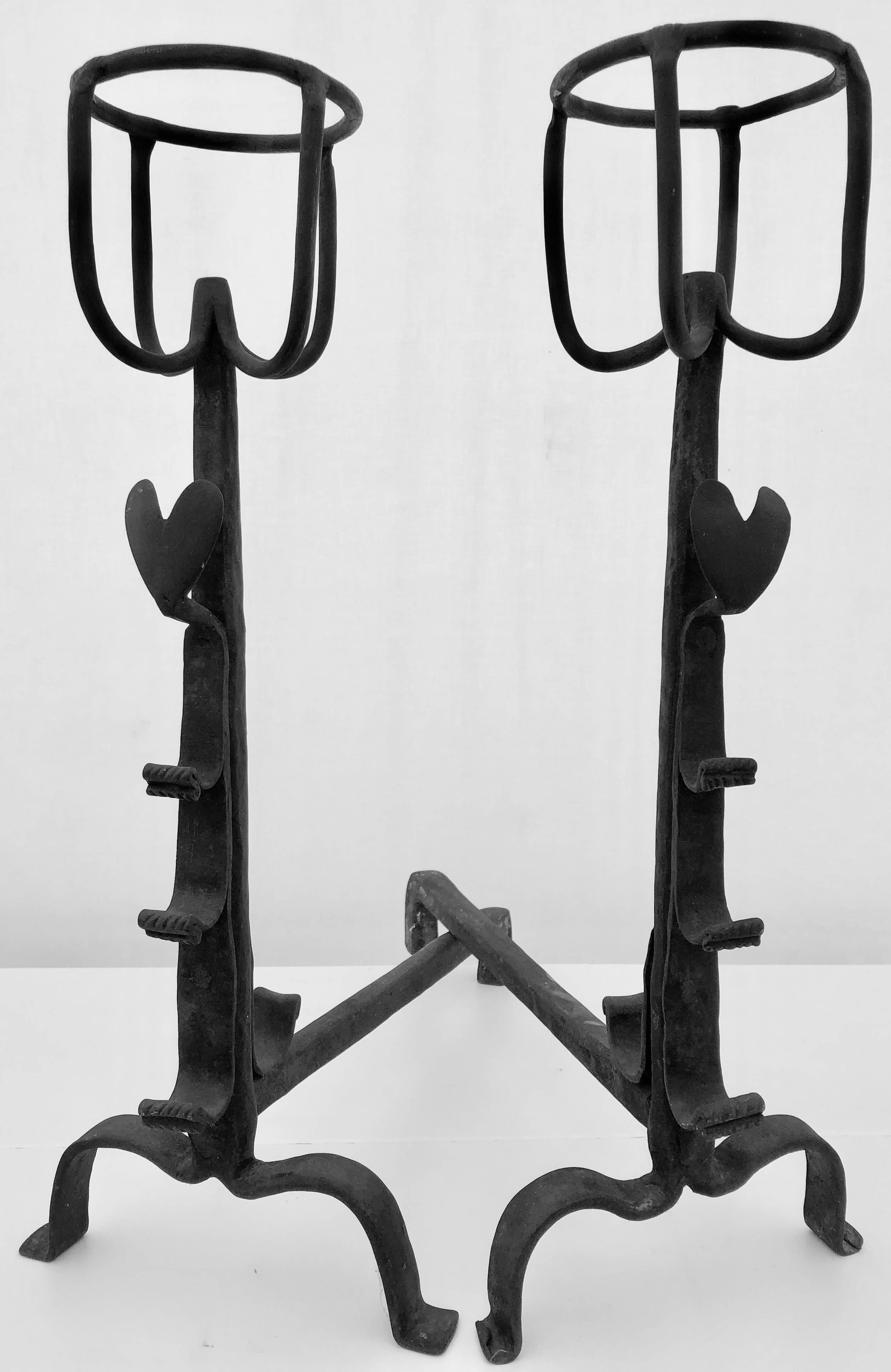 This is a beautiful pair of French early 18th century andirons hand made by a blacksmith in solid forged iron and enhanced by two hearts in the front and topped with two original receptacles, used to keep food or liquids warm in a fire. There are