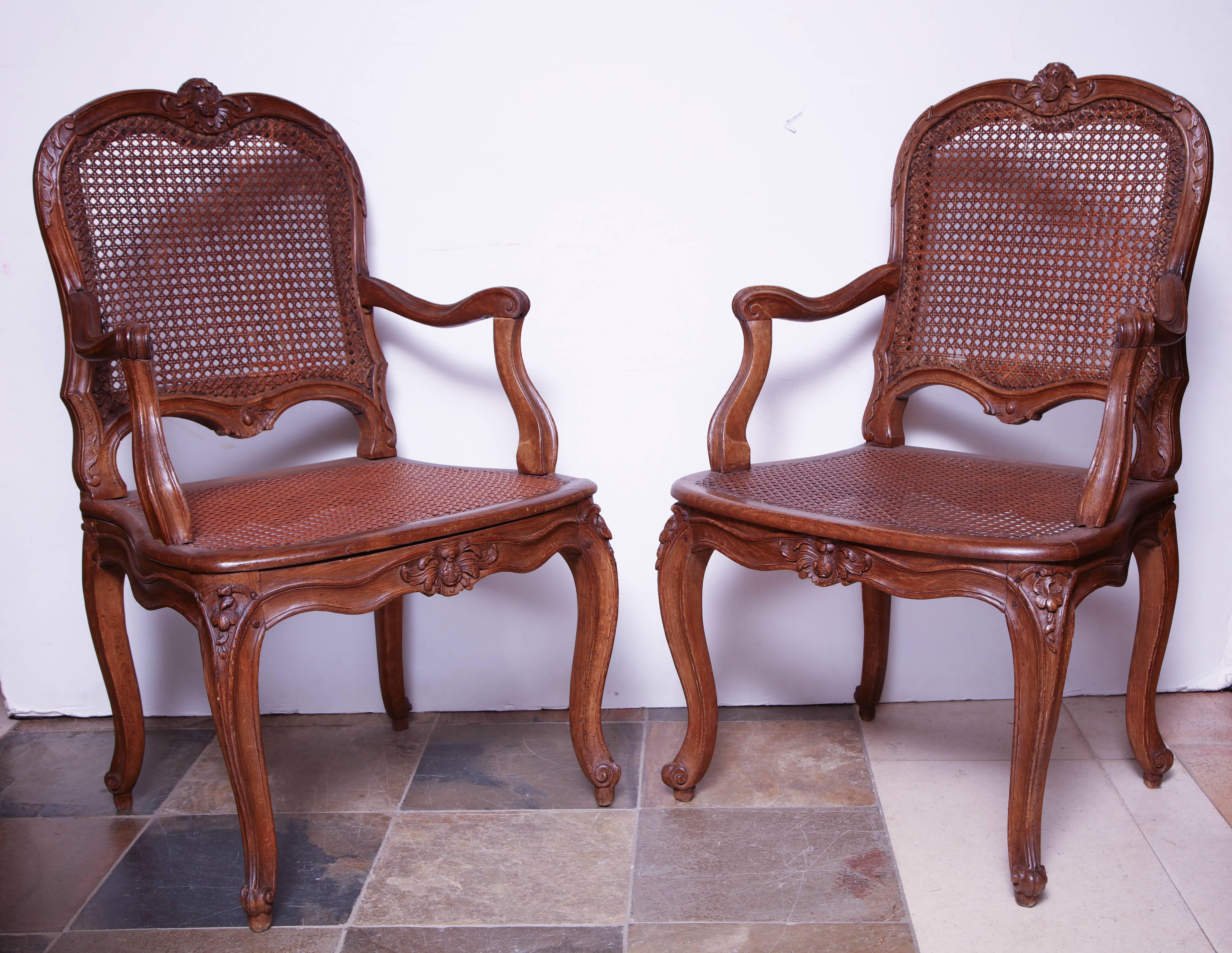 Pair of Louis XV carved Provincial Fauteil's with caned seats and backrests with silk upholstered cushions, carved crests, rails, arms, and legs.