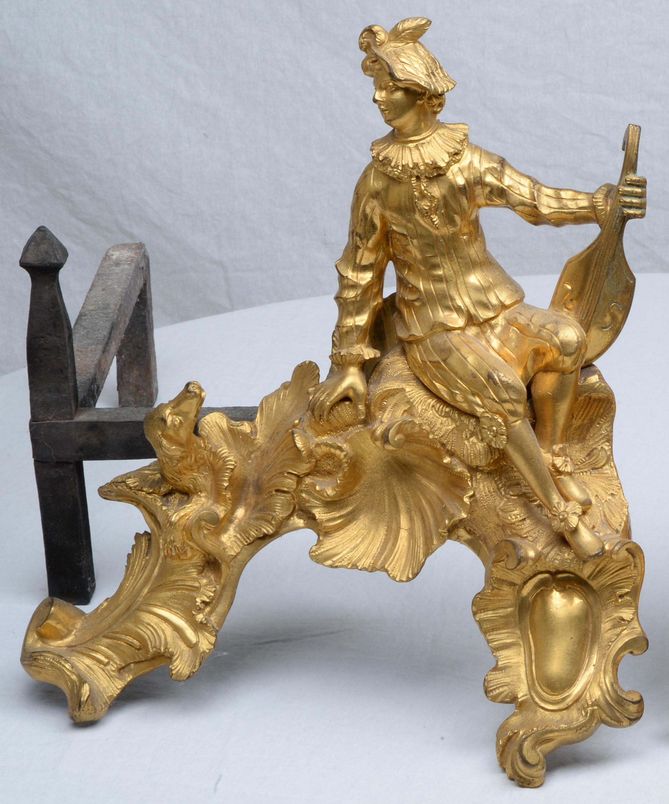 Pair of Louis XV Ormolu Andirons, depicting a seated gallant with a guitar and his companion, each with a dog looking up and rocaille scrolling. with log rests (dimensions include the log rests).

With their asymmetrical forms, the scrolling foliage