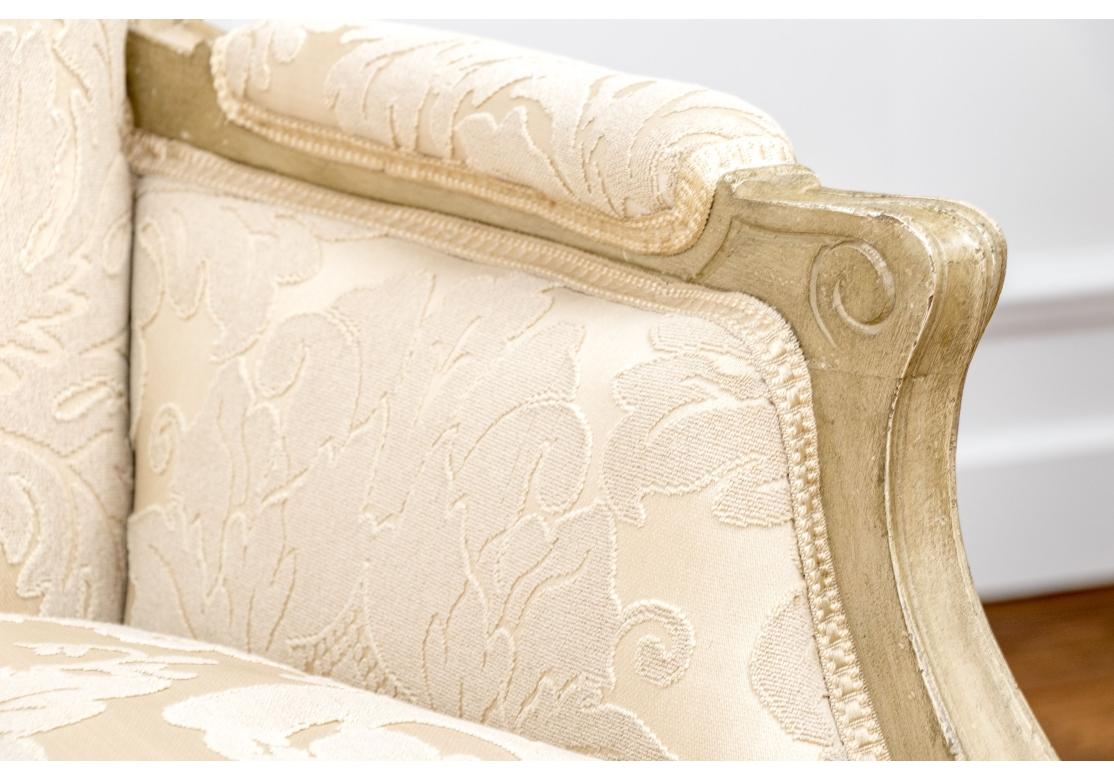 Elegantes bergeres de tamaño amplio con decoración tallada pintada de color topo pálido. Con barandillas de cresta tallada en forma foliada y extremos de los brazos acanalados. El faldón de riel con centro floral, elevado sobre patas cabriolé con